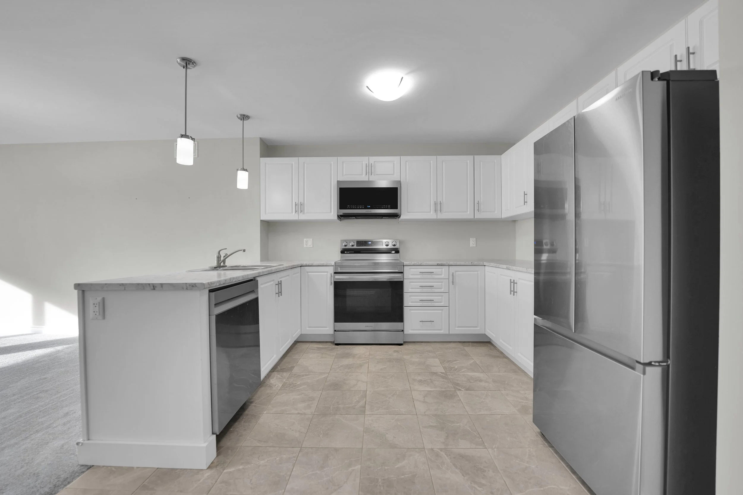 Modern kitchen with white cabinets, stainless steel appliances including a stove, microwave, and refrigerator, beige tiled floor, and two pendant lights hanging from the ceiling.