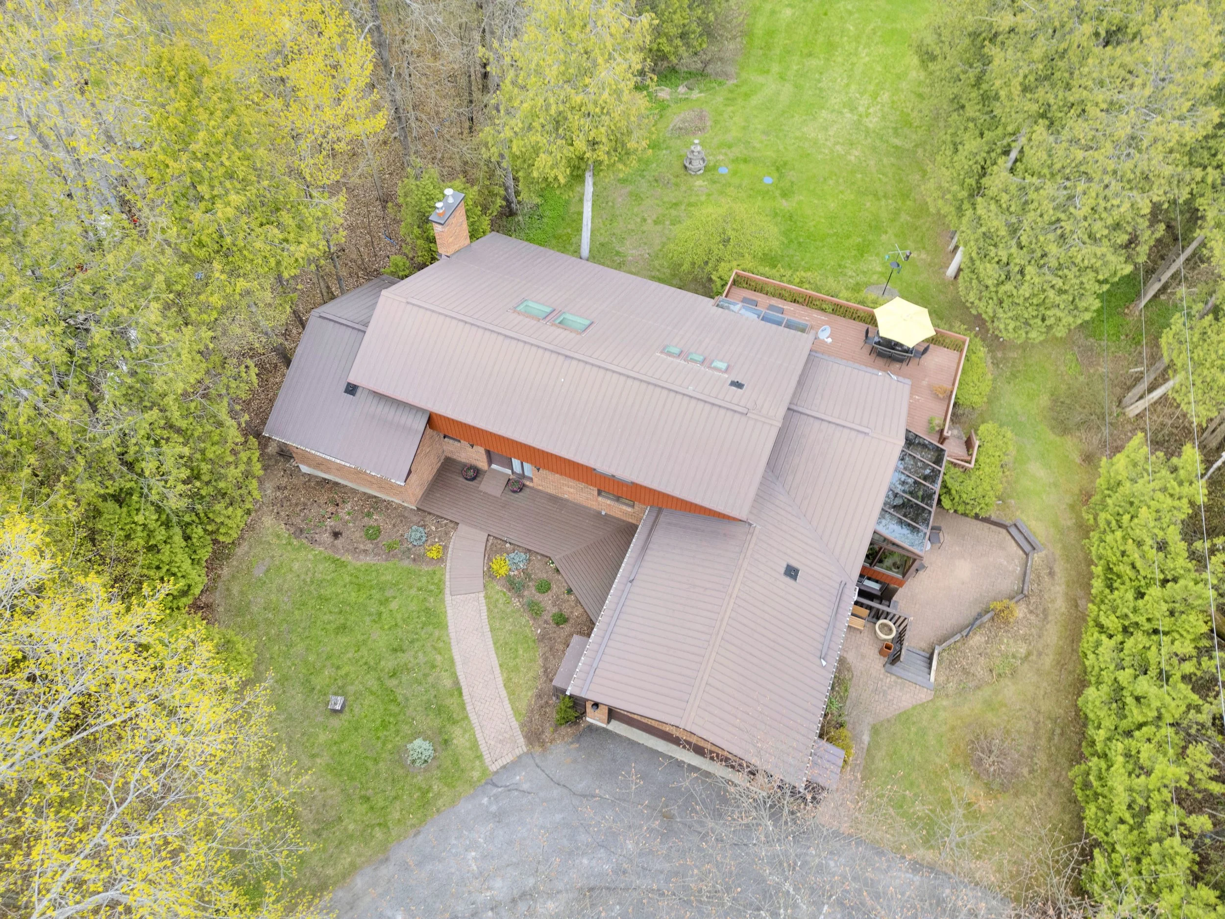 Ottawa Real Estate Media Services  Aerial view of a house with a brown roof surrounded by green trees and a well-maintained lawn. There is a wooden deck with outdoor furniture and a yellow umbrella, and a paved walkway leading to the entrance.
