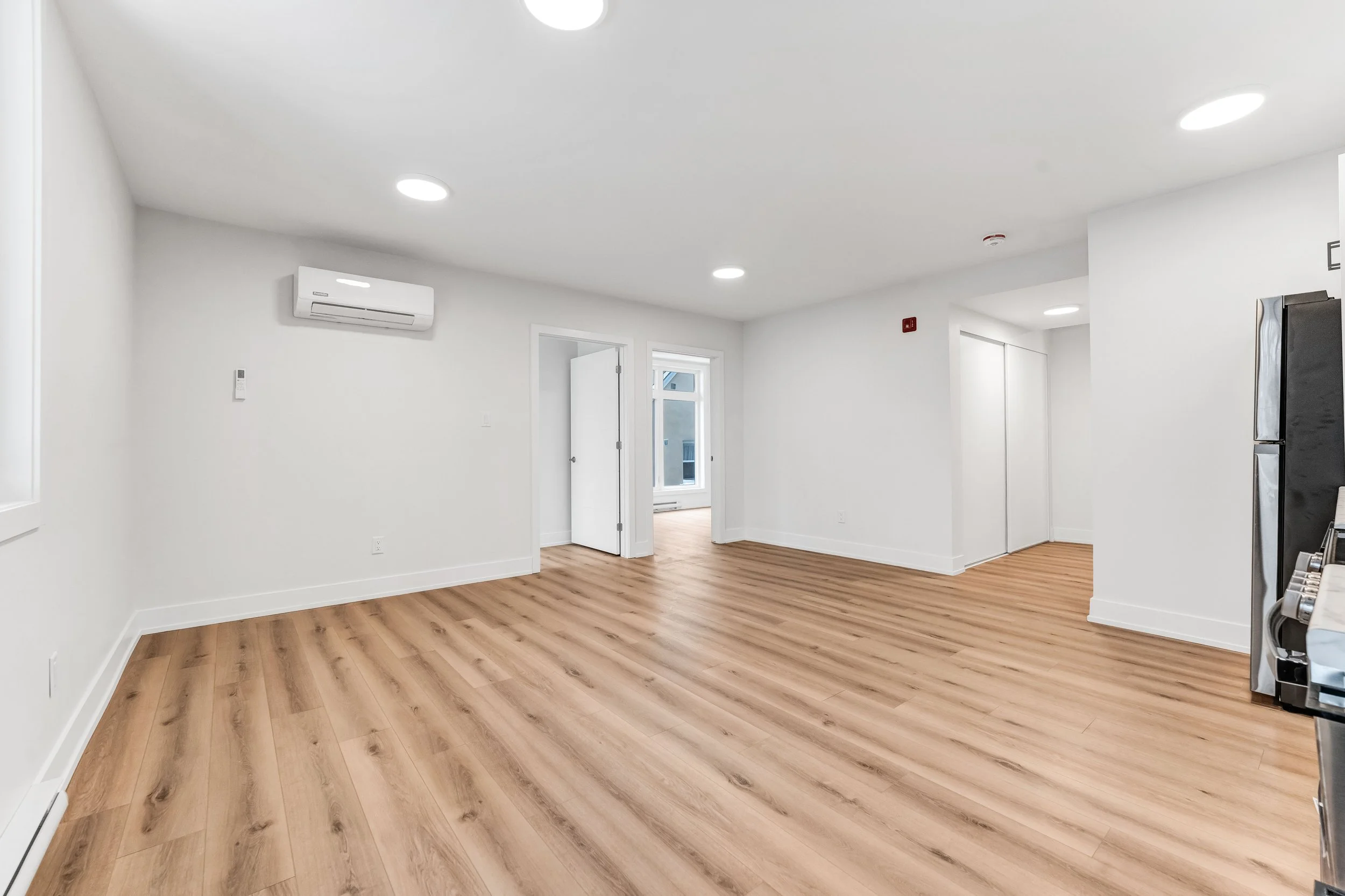 Empty room with light wood flooring, white walls, ceiling lights, and a window.