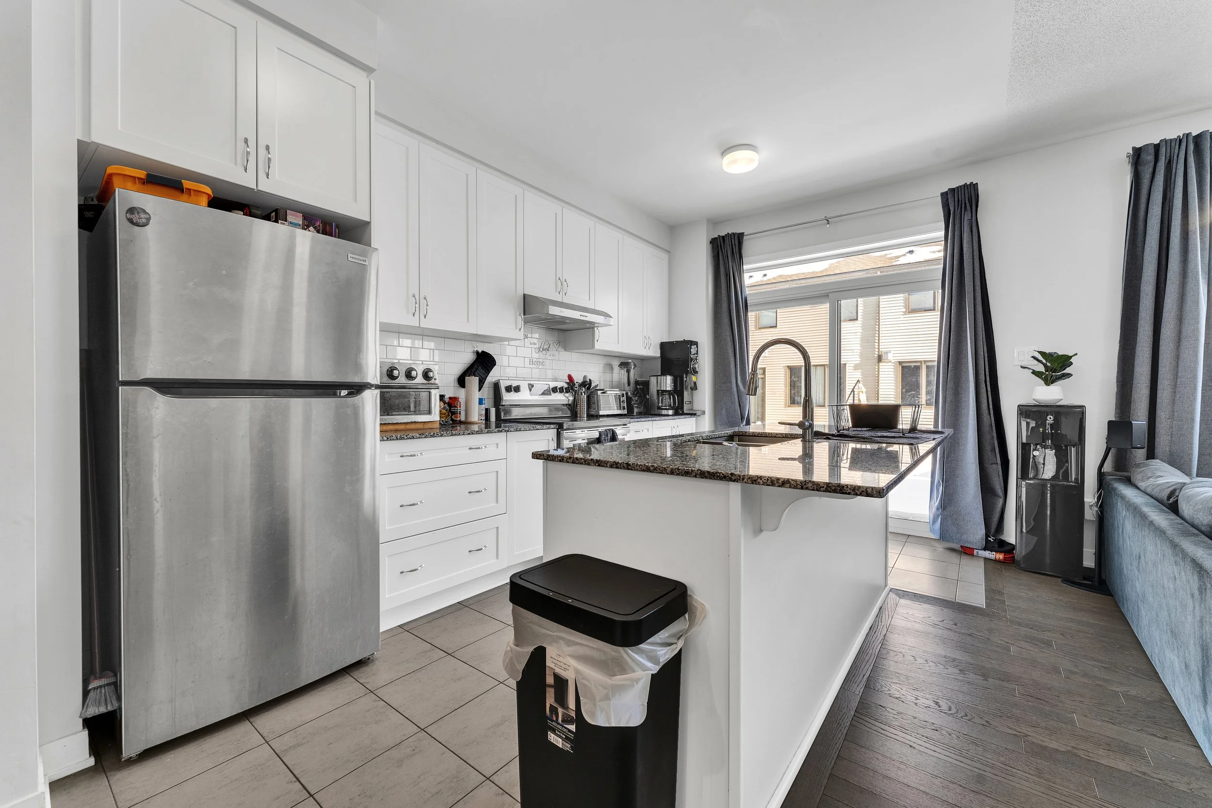 Kitchen with white cabinets, granite countertop island, stainless steel refrigerator, and various small appliances, with a living room area in the background.