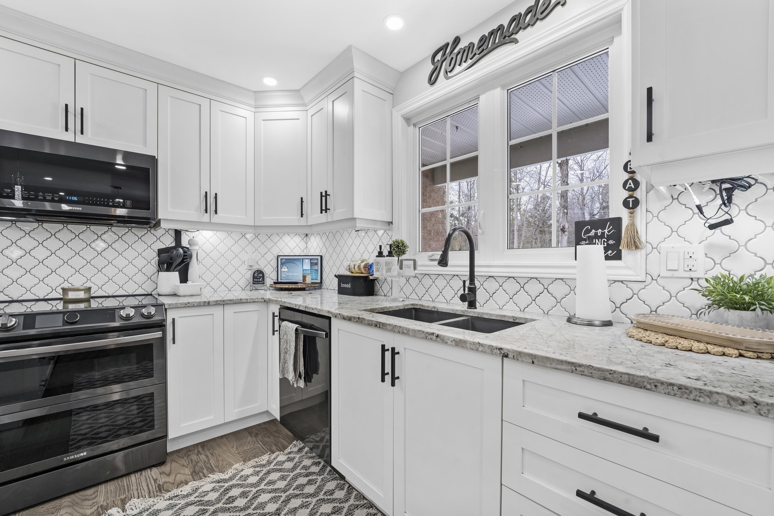 Ottawa RealModern kitchen with white cabinetry, granite countertops, stainless steel appliances, and a patterned backsplash. There is a window above the sink with a decorative sign that reads 'Home,' and various small decorative items on the counter.