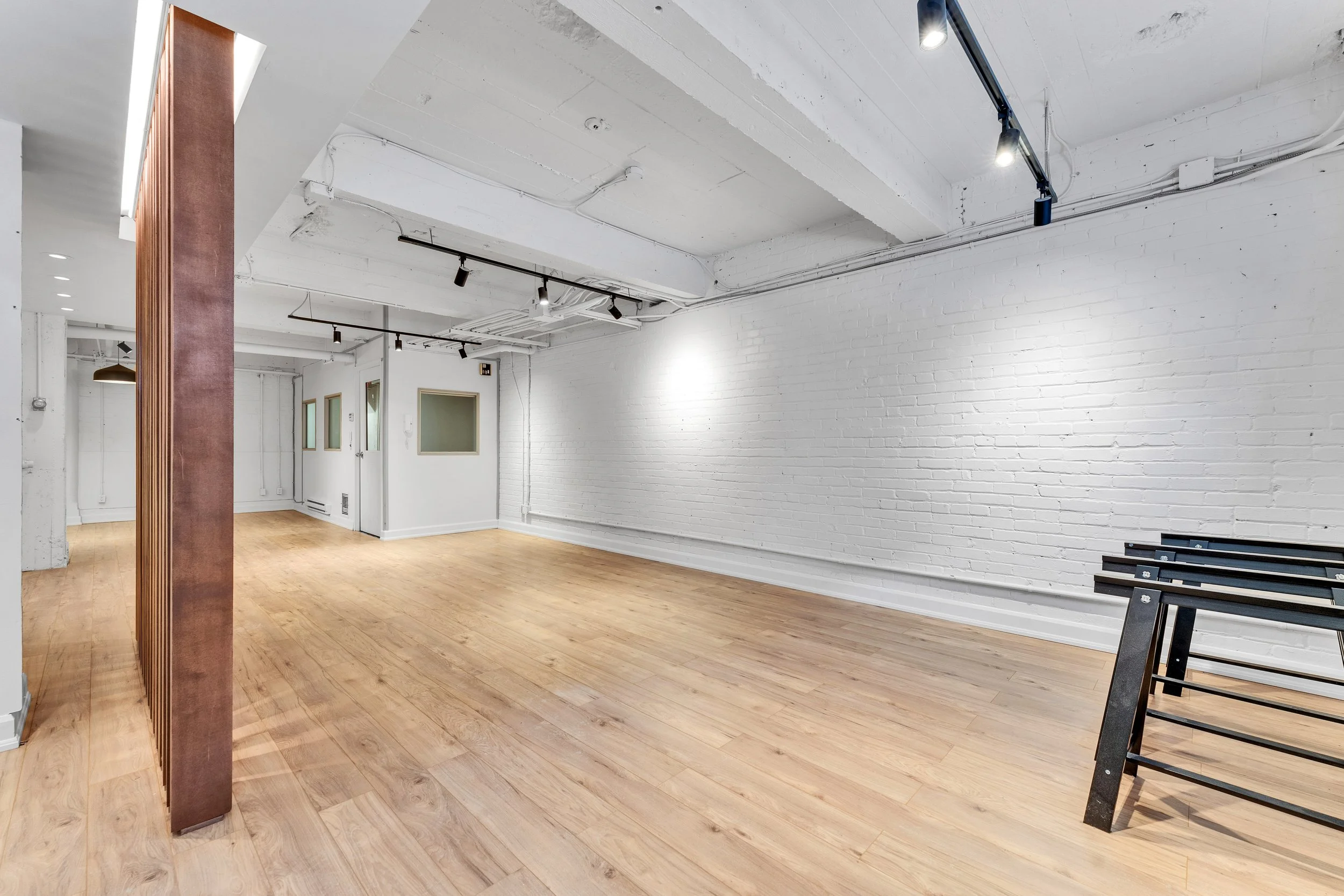 Empty commercial space with white brick walls, light wood flooring, and exposed ceiling with track lighting.