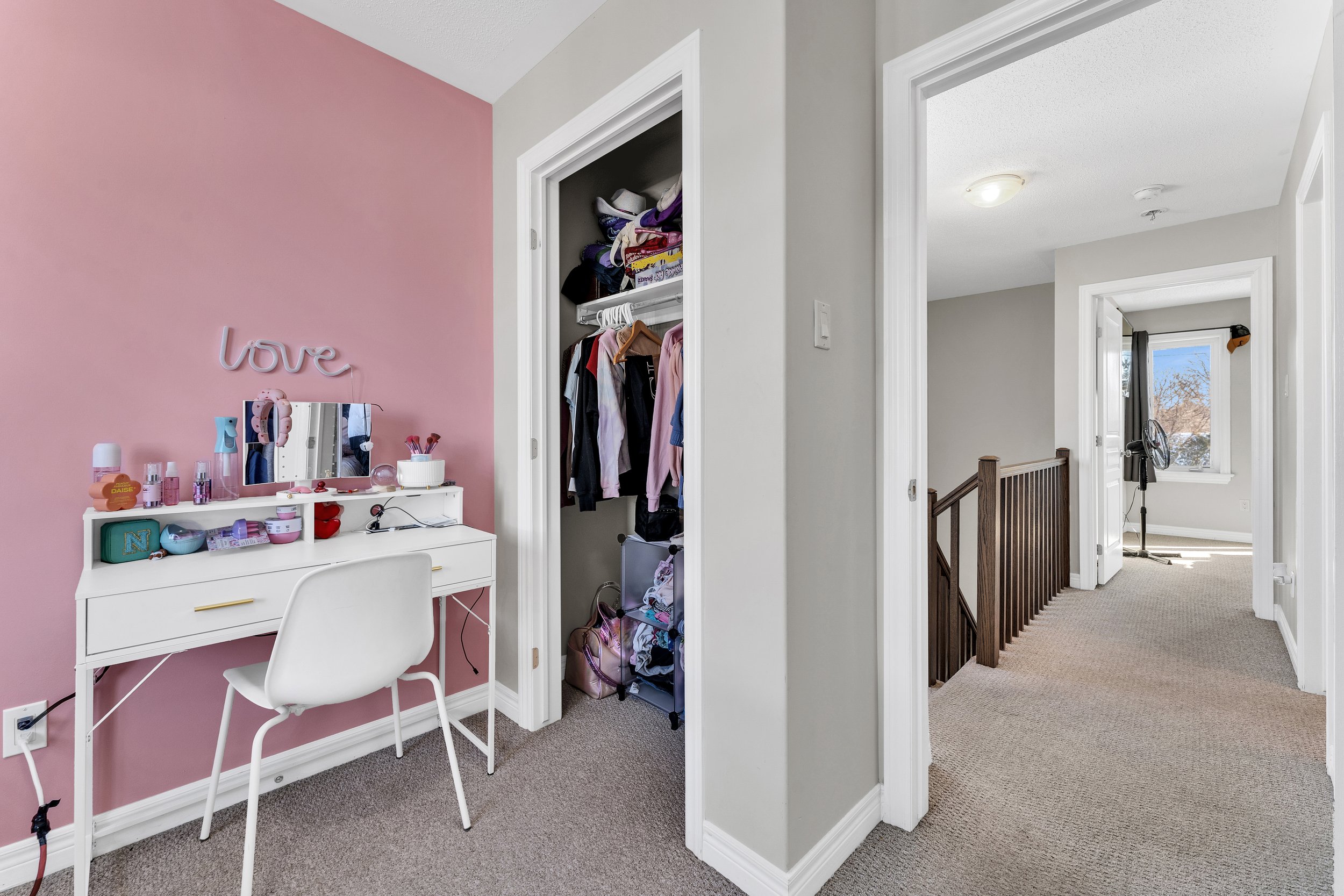 A hallway with a pink accent wall on the left, a white desk with beauty products, a mirror, and a chair in front of it. An open closet with clothes and storage bins is next to the desk. Further down is a staircase with a wooden banister and a landing