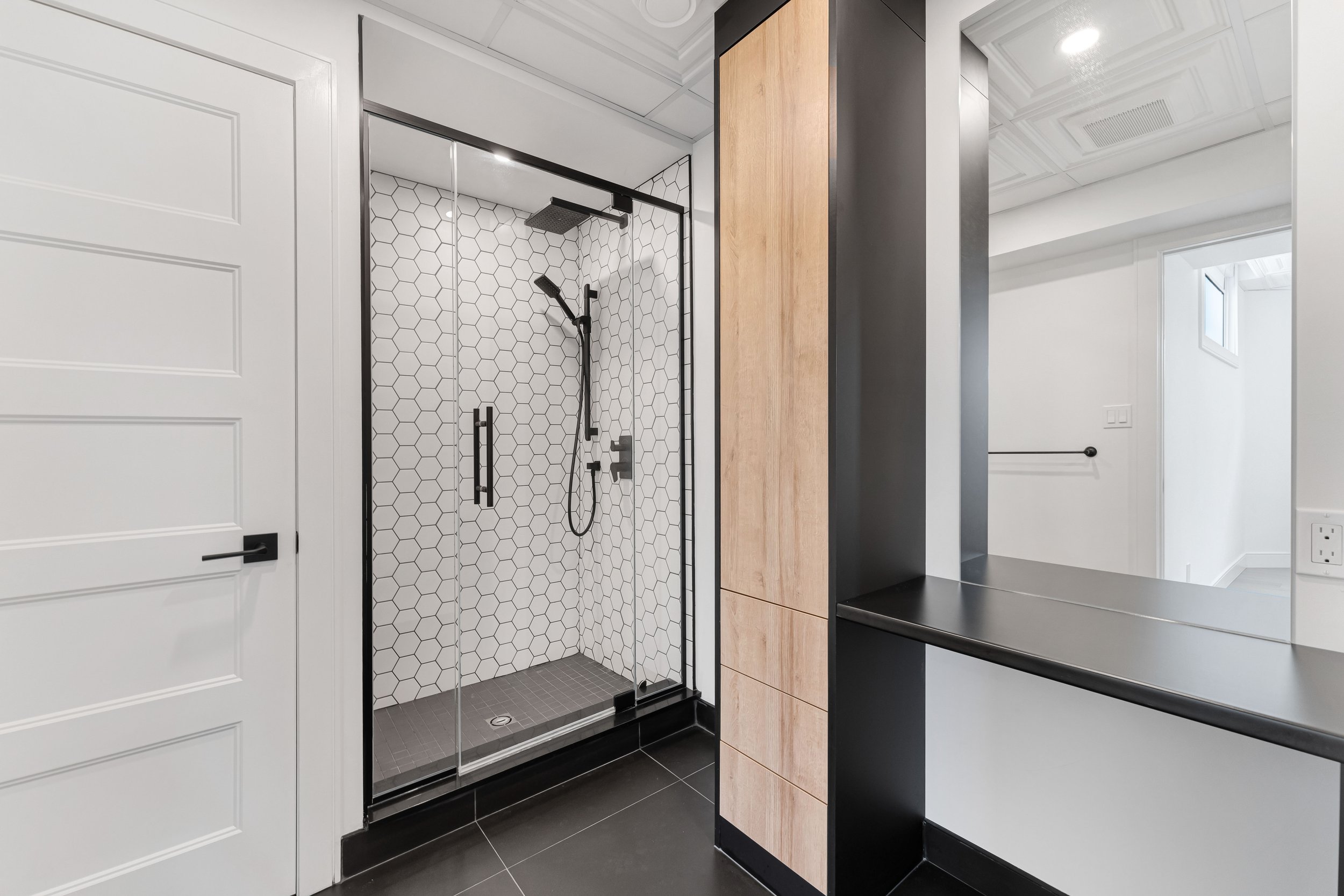 Modern bathroom with glass-enclosed shower featuring hexagonal tile and black fixtures, white panel door, light wood cabinet, and black countertop.