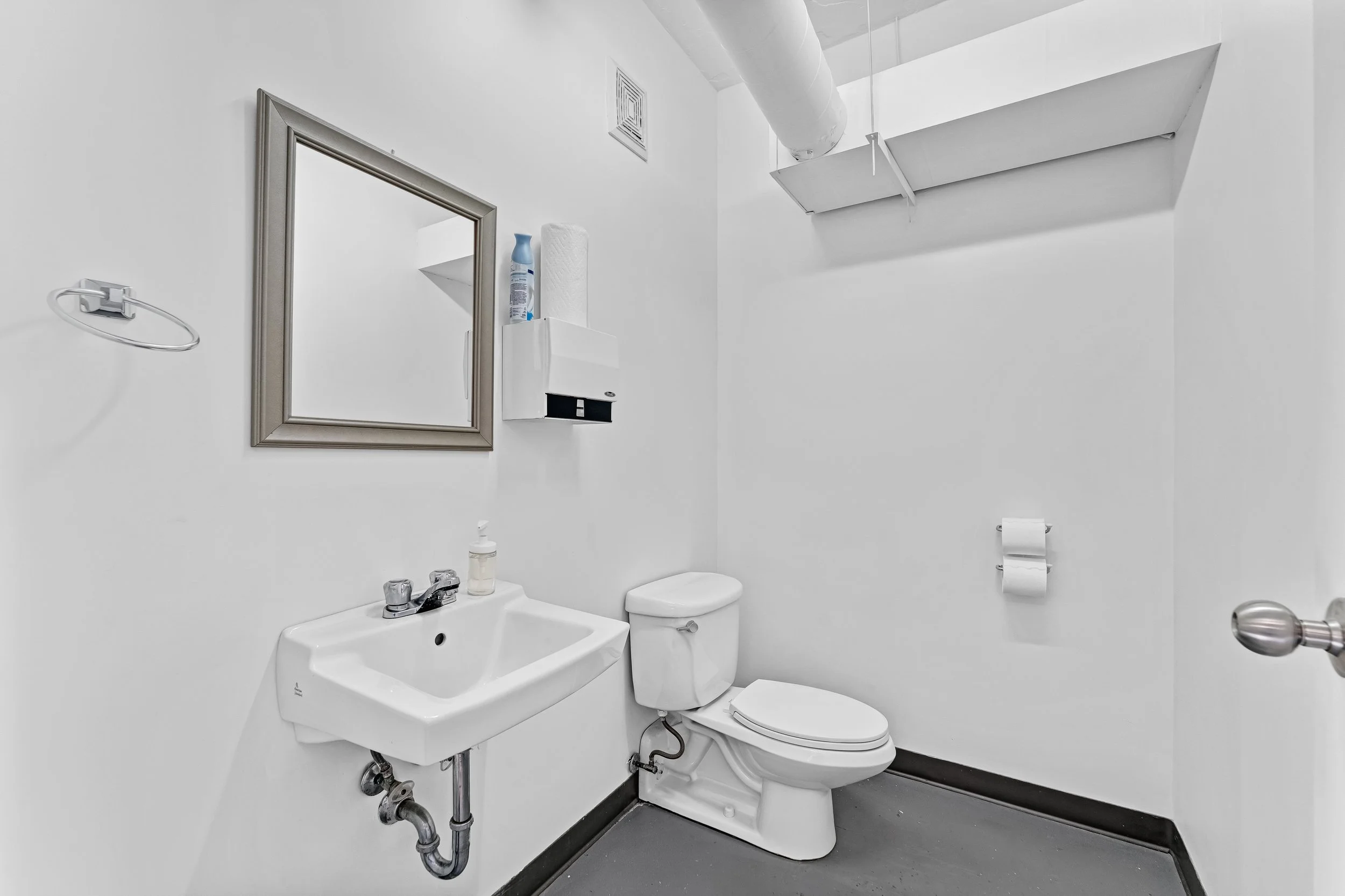 A small, white bathroom with a wall-mounted sink, a toilet, a mirror, a paper towel dispenser, and a roll of toilet paper. There is a ventilation duct on the ceiling.