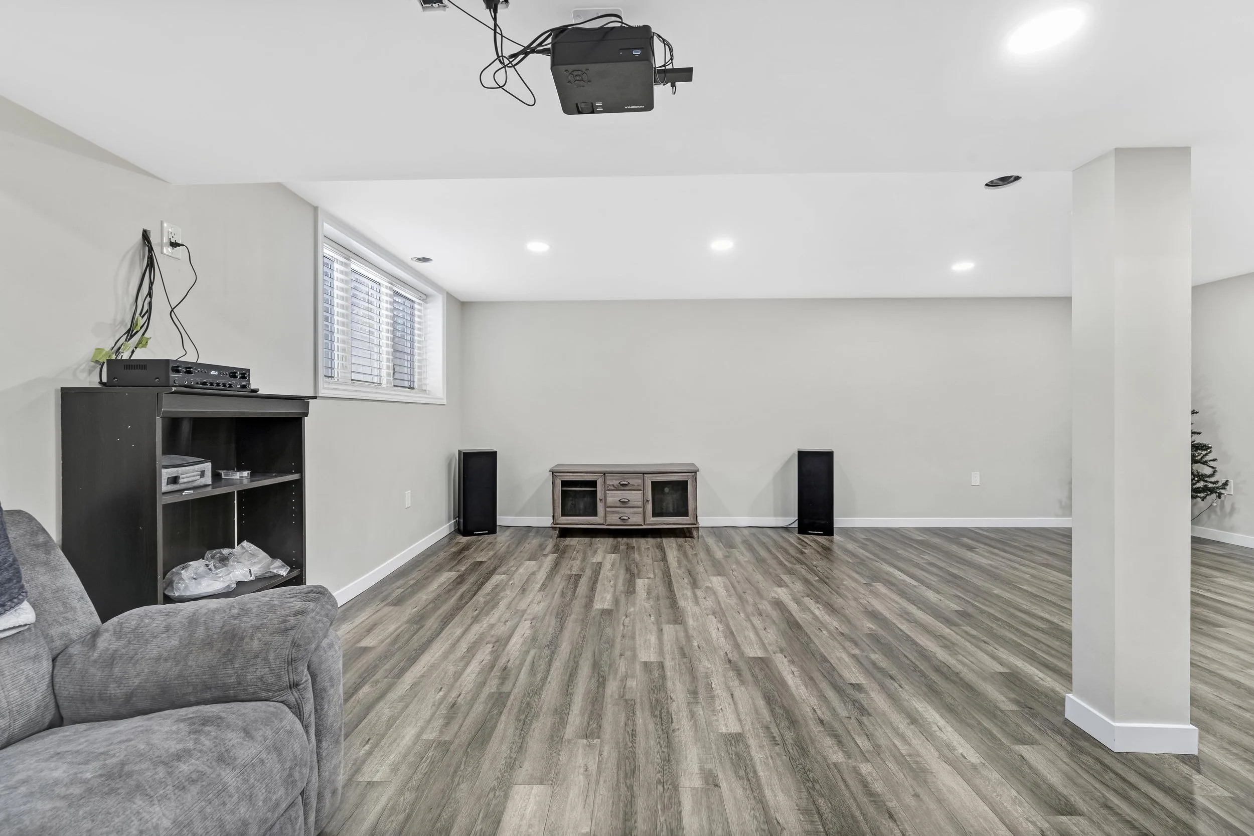 Empty entertainment room with wooden floor, a window with blinds, black speakers on either side of a wooden TV stand, a black shelf unit with electronic devices, and a partially visible gray couch on the left, with a small Christmas tree on the far r