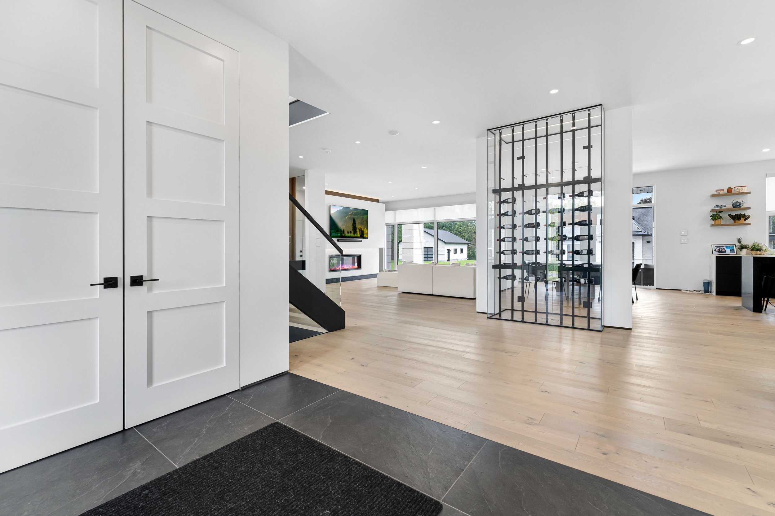 Modern open-concept living area with white walls, light wood flooring, a large wine rack, and a view of the backyard through large windows. The space includes a staircase, a wall-mounted TV above a fireplace, and decor shelves with plants.