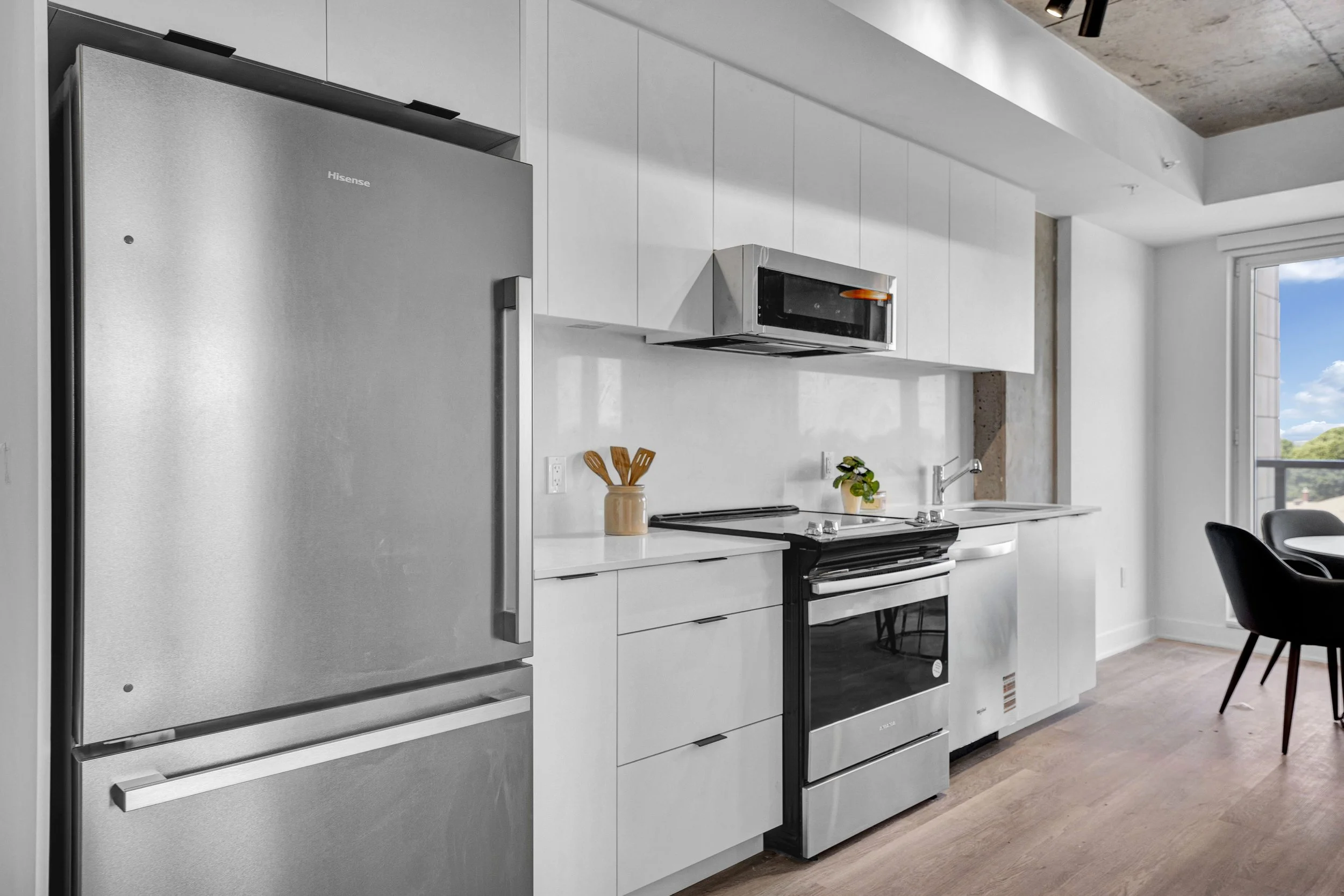 Modern kitchen with white cabinets, stainless steel refrigerator, microwave, stove, and small potted plant on the counter near a large window.