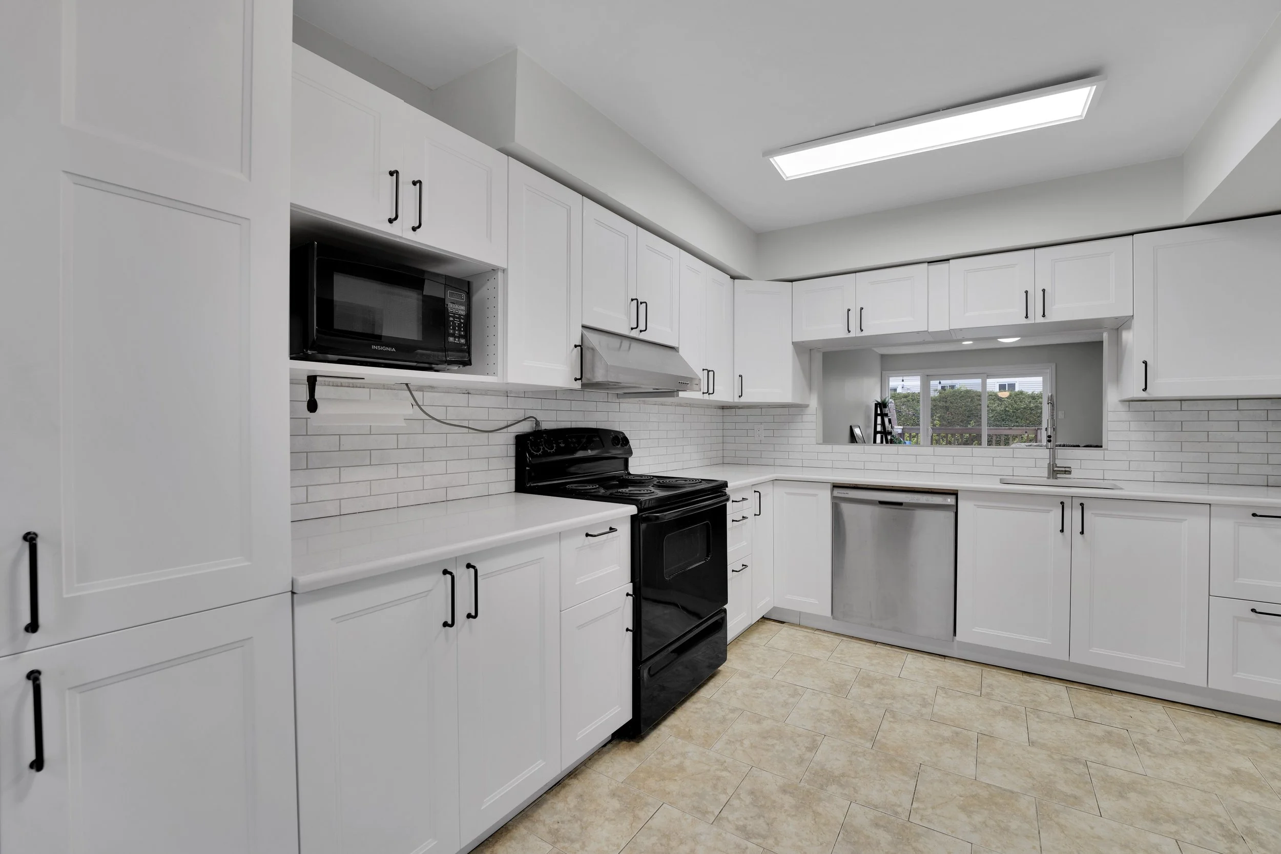 A modern kitchen with white cabinets, black handles, black appliances, a window with a view of greenery, and beige tiled flooring.