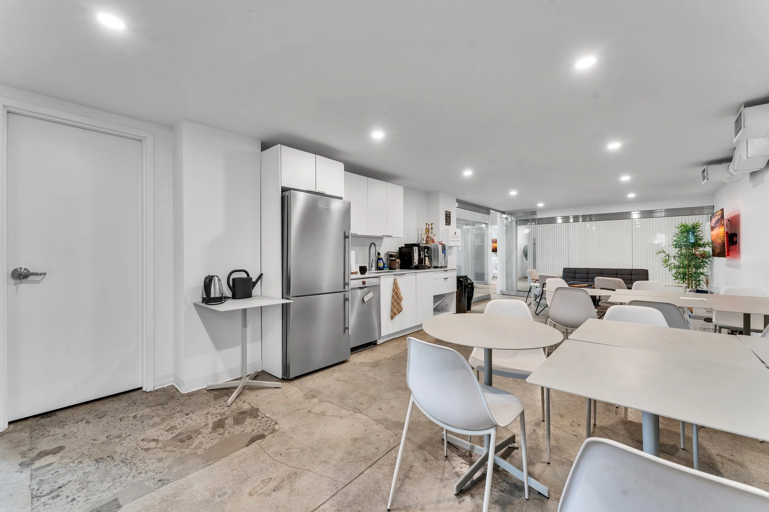 A modern communal kitchen and dining area with white walls and ceiling, concrete flooring, stainless steel refrigerator, white cabinets, and white tables with chairs. There is a sitting area in the background with a large window, a sofa, a potted pla