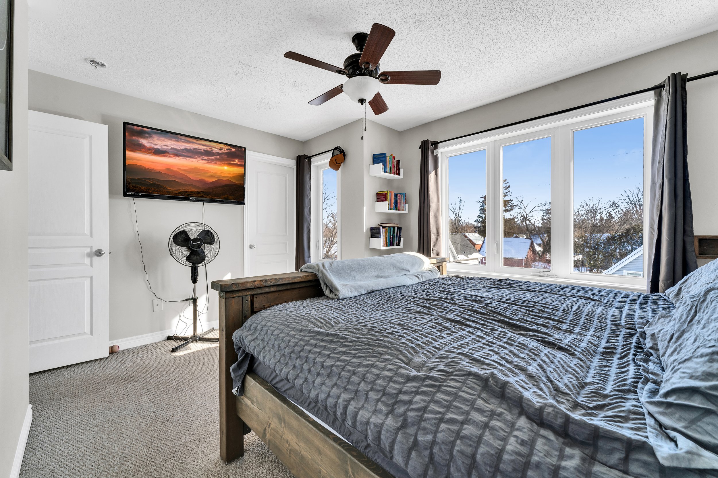 A bedroom with a large bed covered with a gray quilt, positioned near a large window showing a wintery outside scene. The room has white walls, a ceiling fan with wooden blades, a wall-mounted TV showing a sunset landscape, and a small space heater.