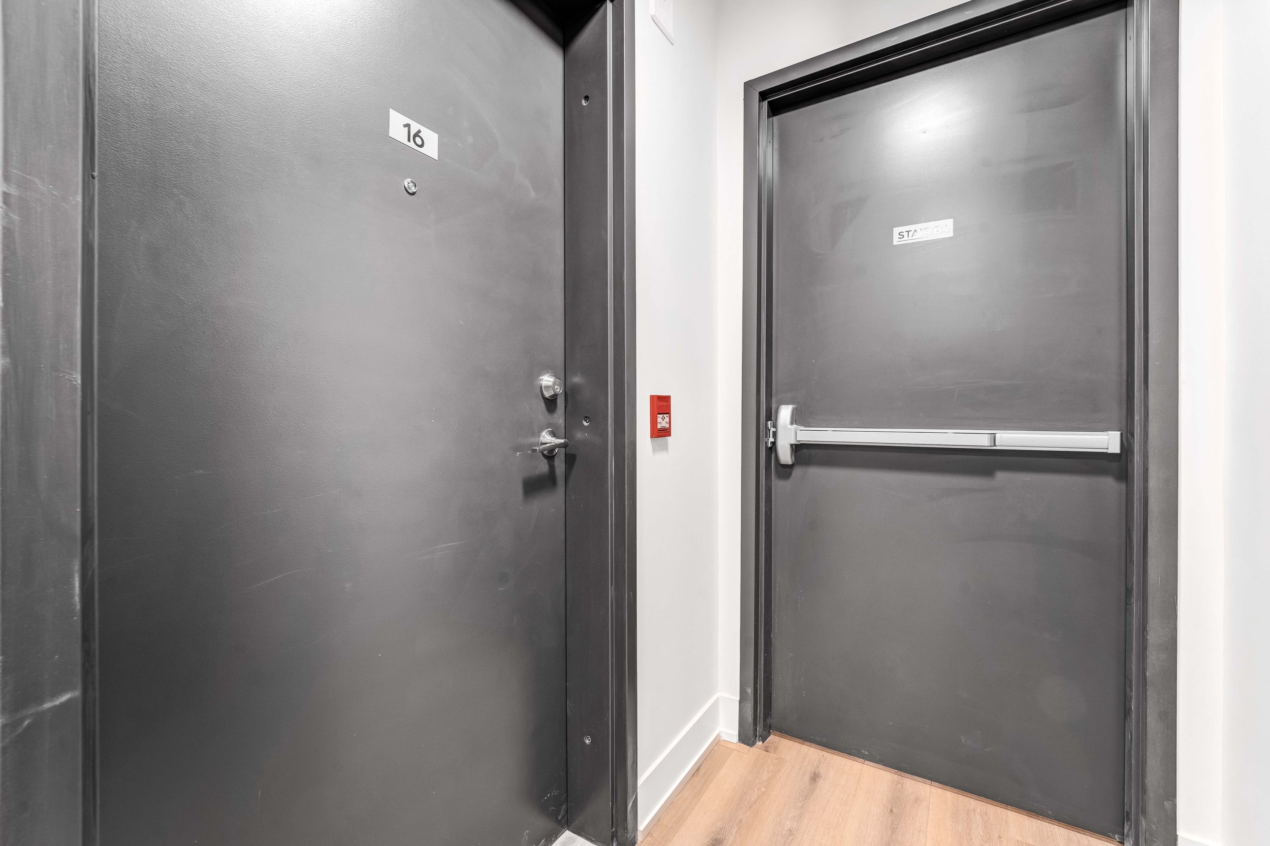 Two dark gray elevator doors, one standard and one fire-resistant, in a building hallway with a fire alarm button on the wall between them. The doors have room numbers 16 and a sign indicating 'STAIR 64'.