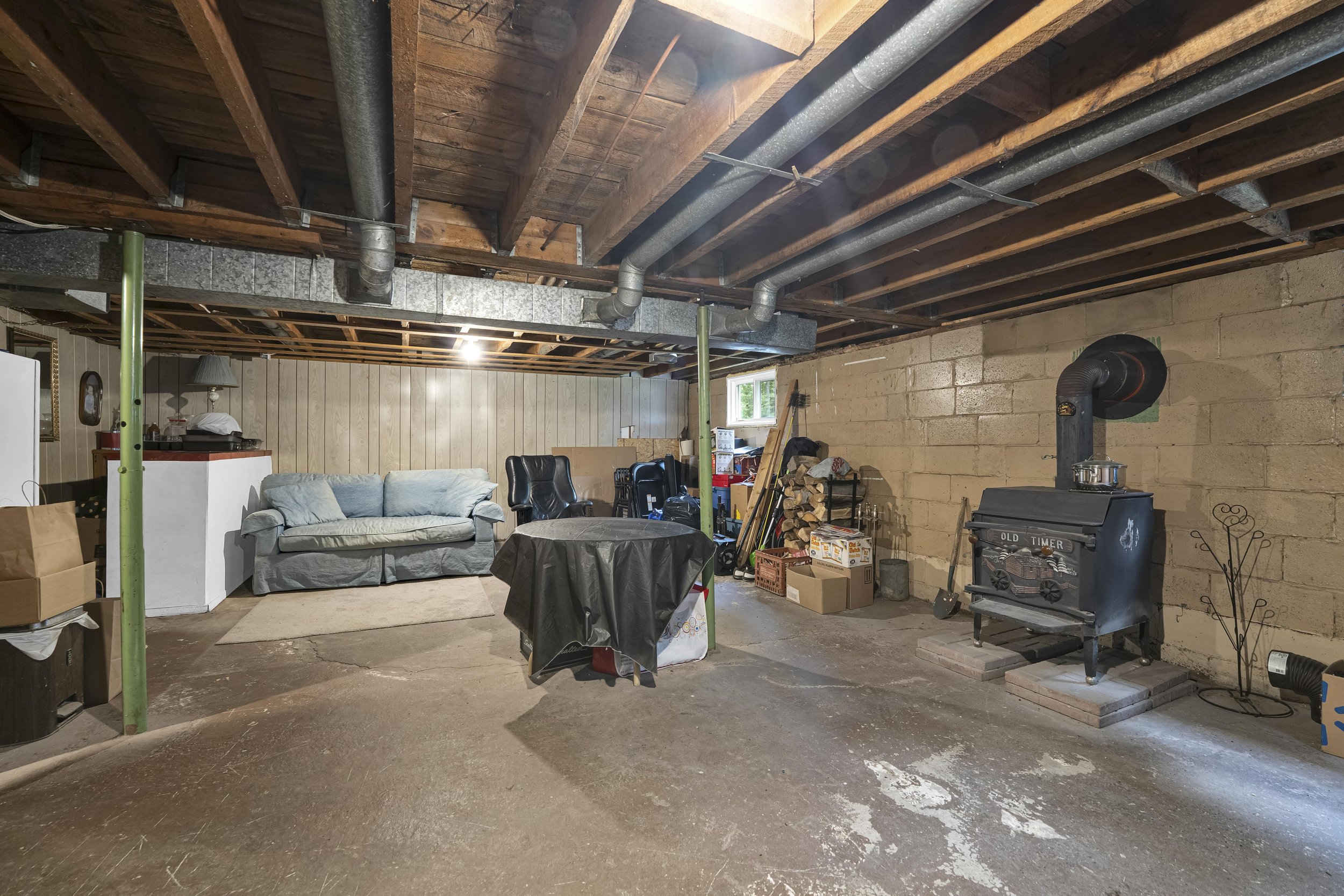 Ottawa Real Estate Photographer A cluttered basement with a couch, boxes, woodpile, wood-burning stove, and miscellaneous items, exposed ceiling beams and pipes, and a small window.