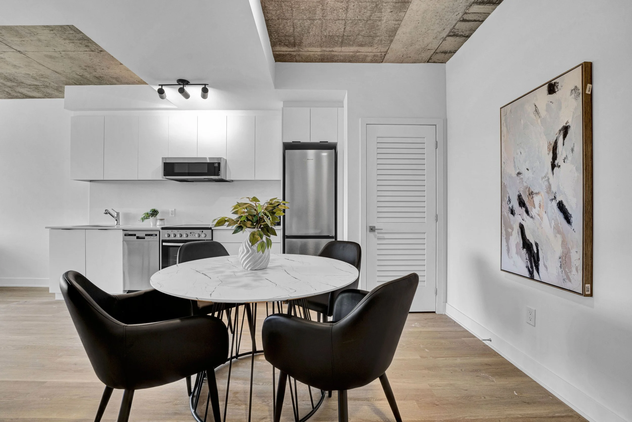 Modern kitchen with white cabinets, stainless steel refrigerator and microwave, black dining chairs around a white marble table, abstract artwork on the wall, wooden flooring, and a ceiling with exposed concrete accents.