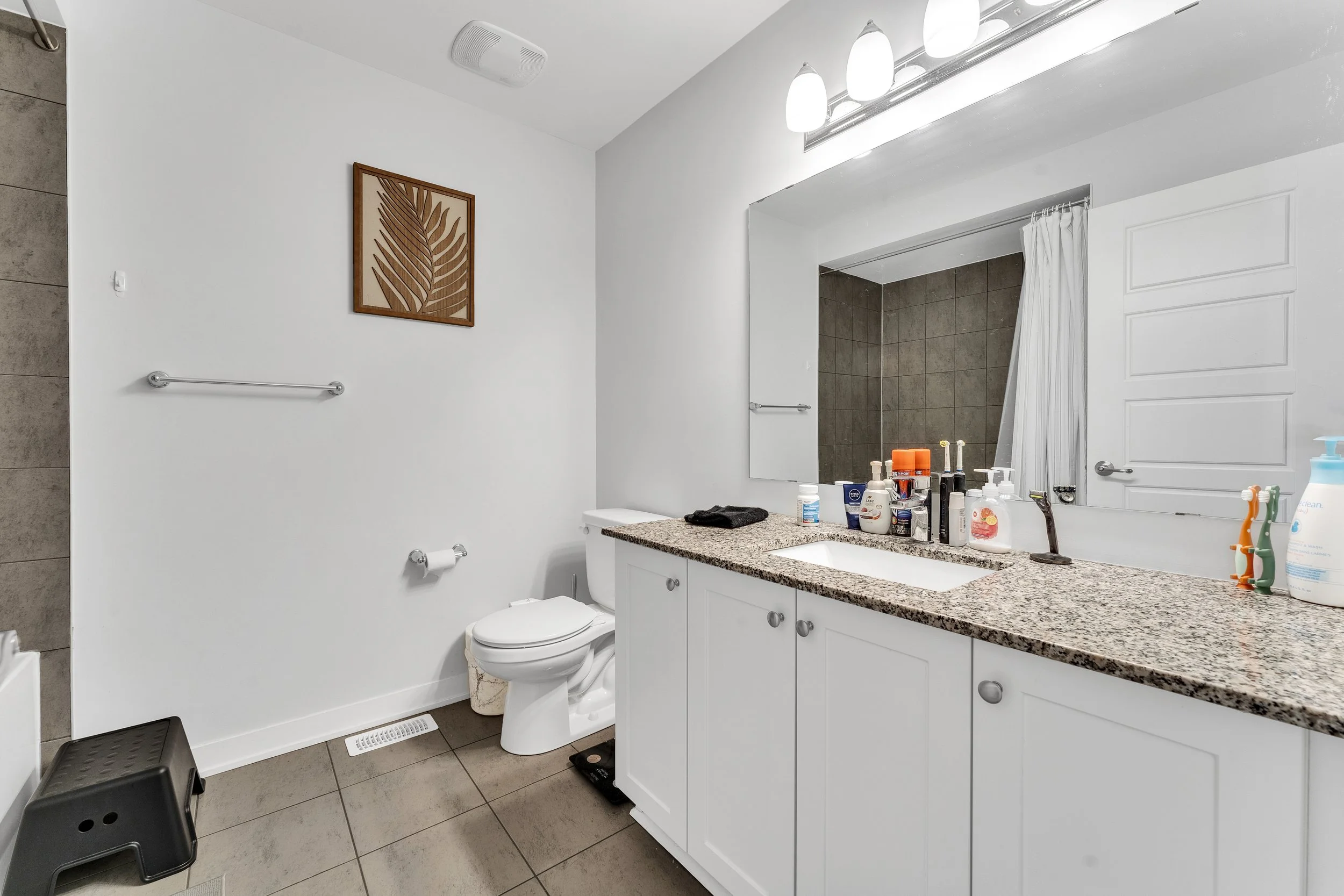 Bathroom with white cabinets, granite countertop, large mirror, toilet, black step stool, decorative wall art, white walls, and tiled shower area with a curtain.