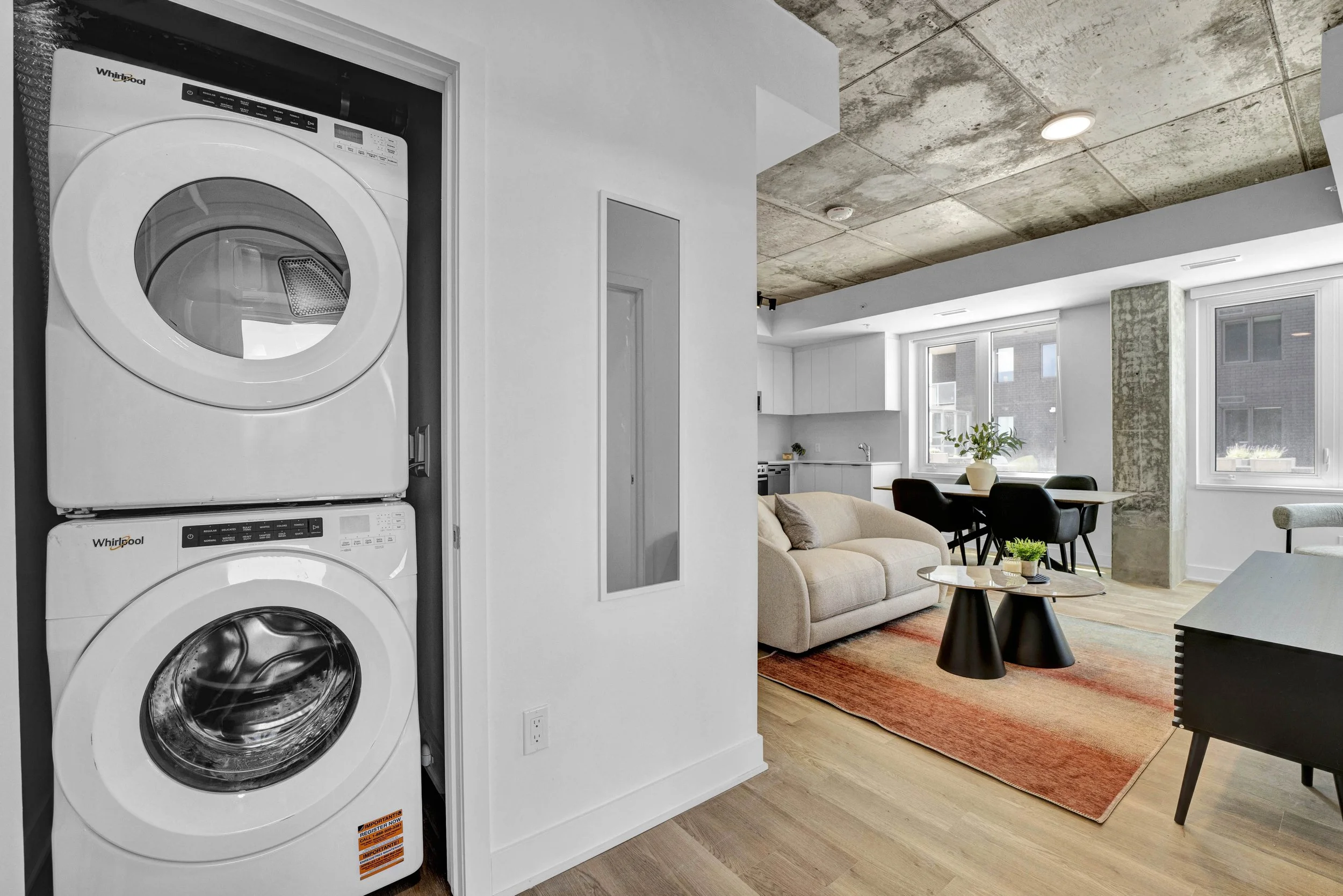 A laundry area with a stacked white washing machine and dryer in a closet next to a white wall in an open-concept living space. The living space has large windows, a beige couch, a black dining table with chairs, a potted plant, and a red and orange 