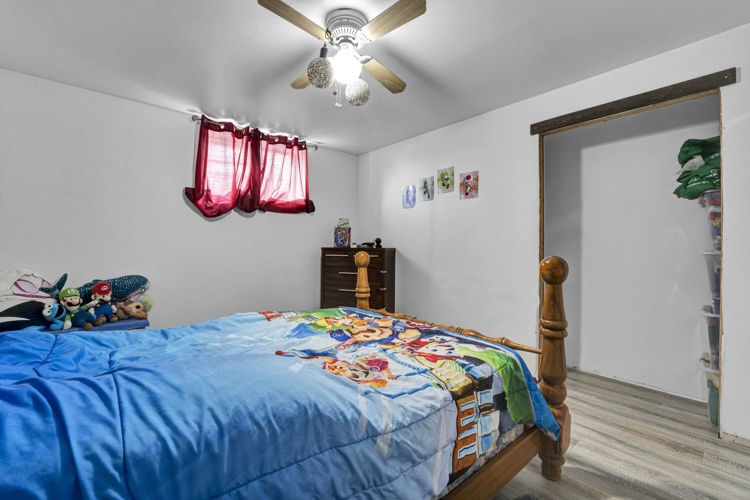 Bedroom with wooden bed frame, blue themed bed with a 'Super Mario' blanket, red curtains, Nintendo character plush toys on the bed, wall decorations of Nintendo characters, small dresser with toys, and a doorway leading to another room.