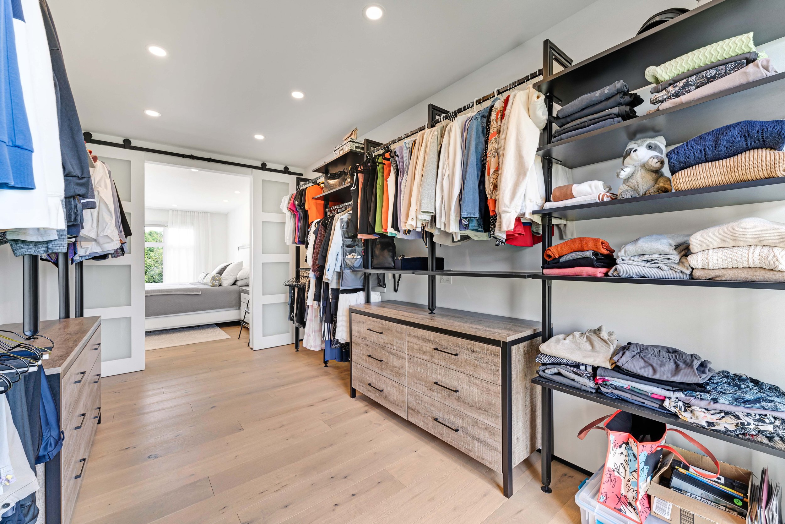 Modern walk-in closet with hanging clothes, shelves with folded garments, a wooden dresser, and sliding barn doors leading to a bedroom with a bed and natural light.