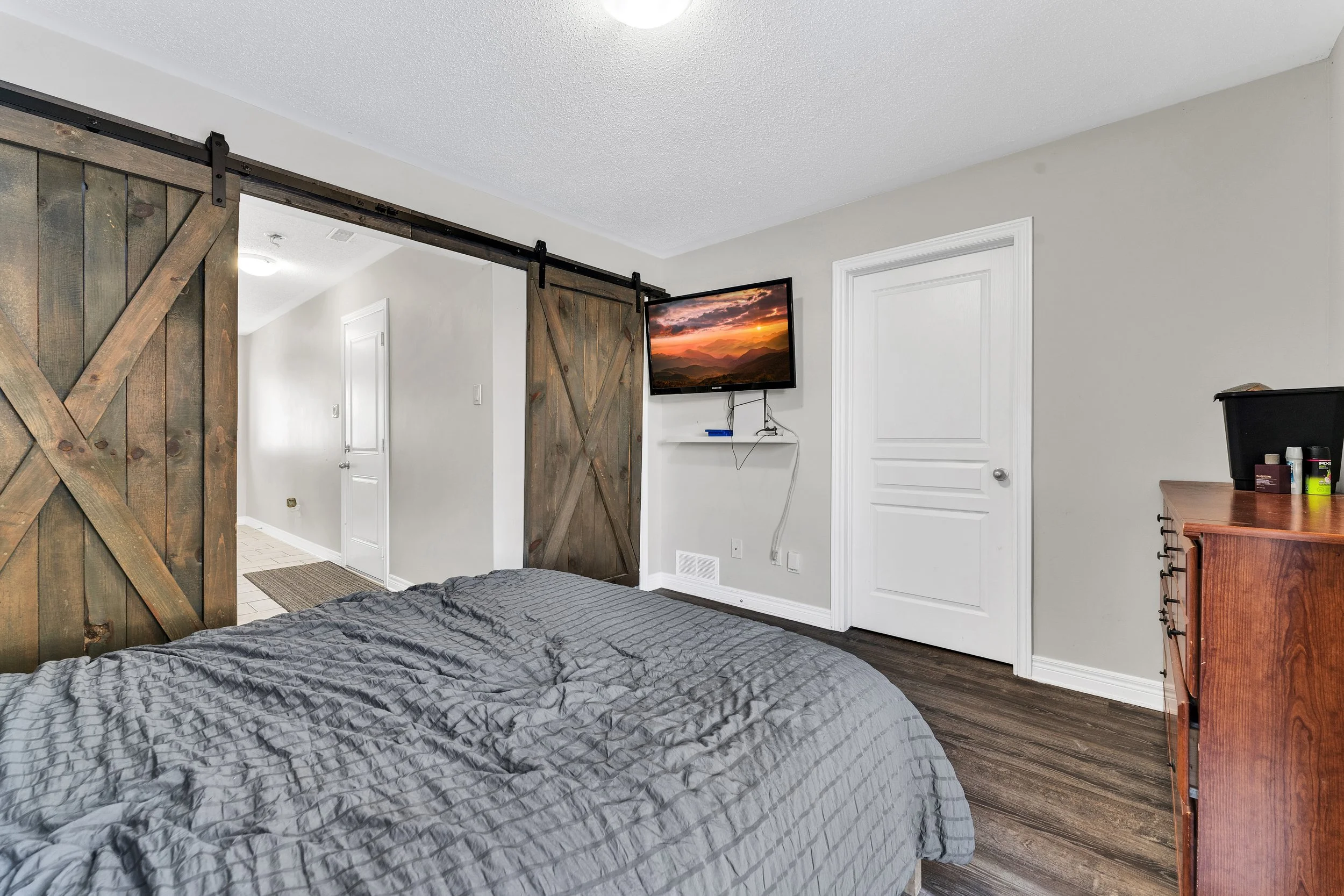 Bedroom with a bed, sliding barn door, wall-mounted TV, white door, wooden dresser, and hardwood flooring.