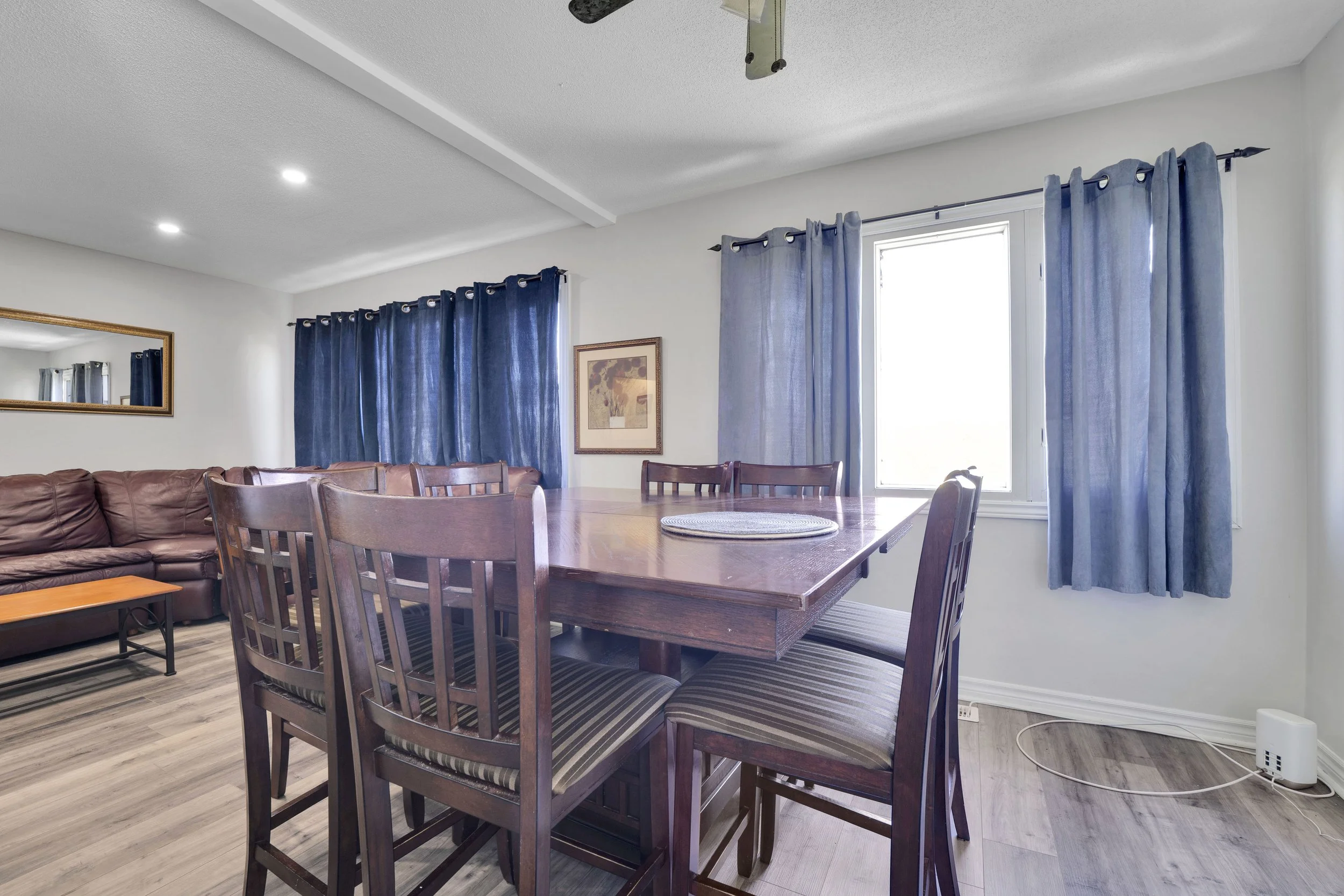 Ottawa Real Estate Media Services  Dining and living room area with a large window covered by blue curtains, a wooden dining table with four chairs, a leather couch, and a mirror on the wall.