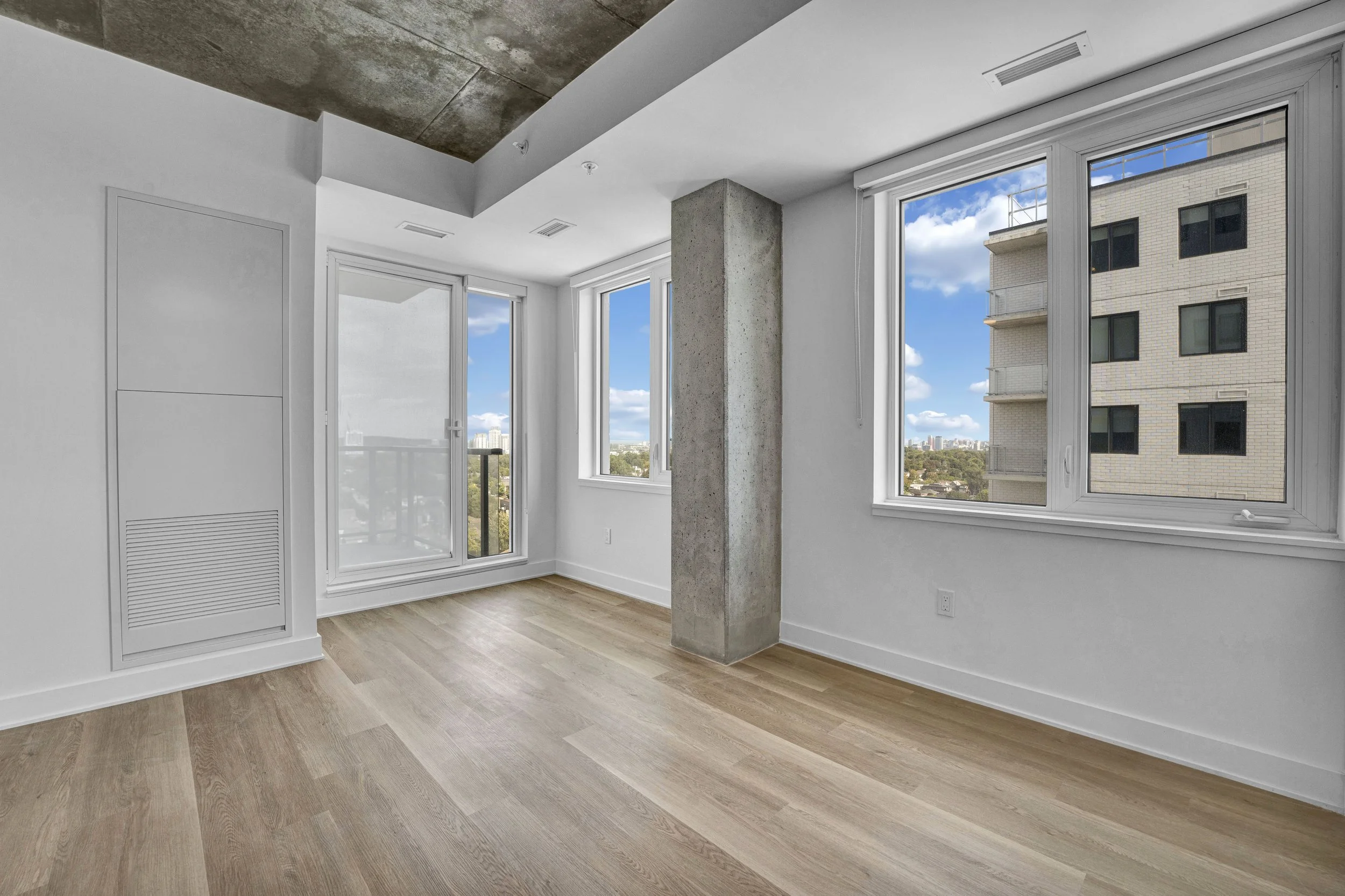 Empty room with large windows showing a cityscape and blue sky, with white walls, light wood flooring, and a concrete column.