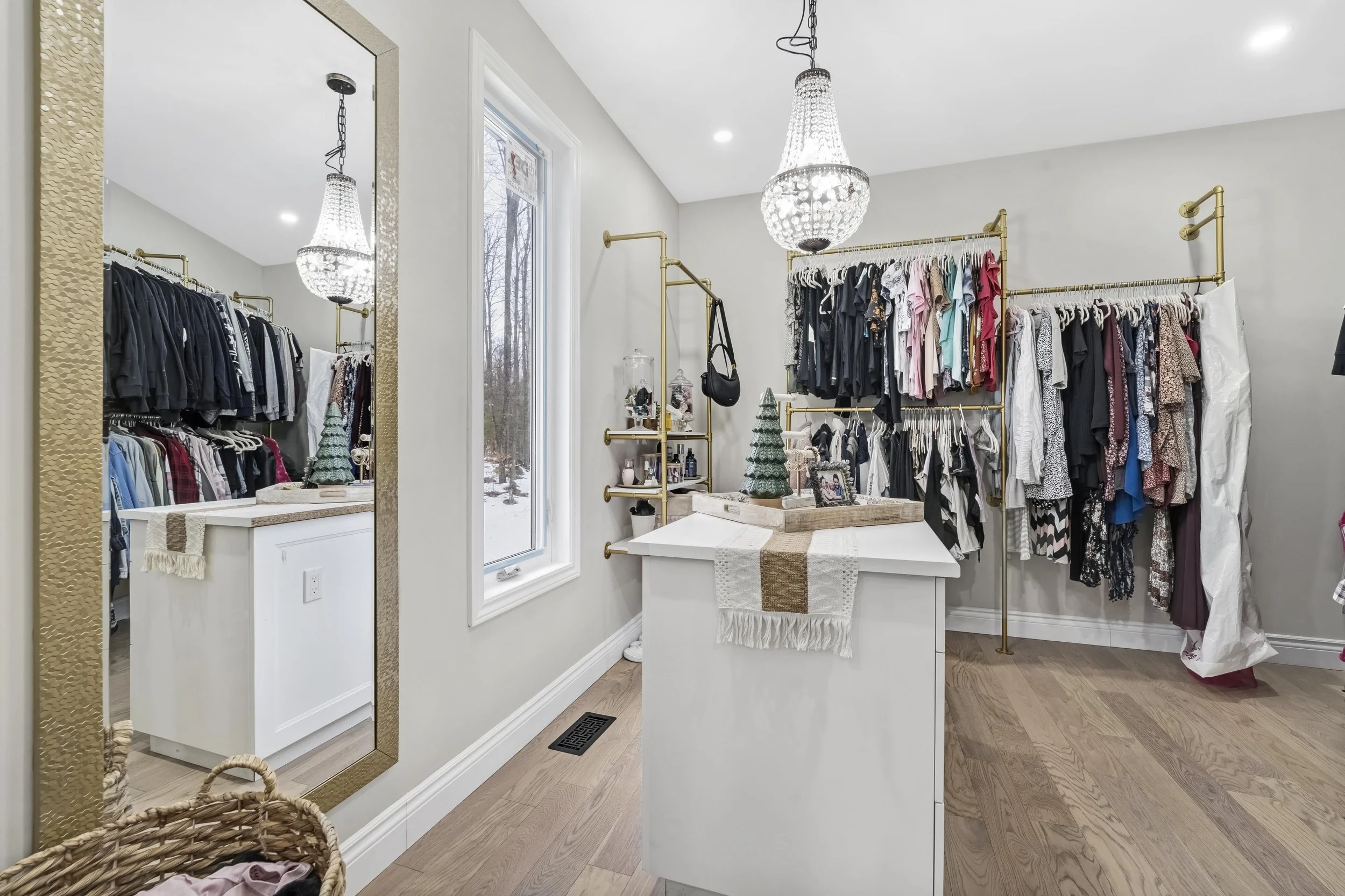 A walk-in closet with clothing racks filled with various clothes, a large mirror on the left wall, a window showing a snowy outdoor scene, a chandelier hanging from the ceiling, white walls, a white storage island with decorative items, and a basket 