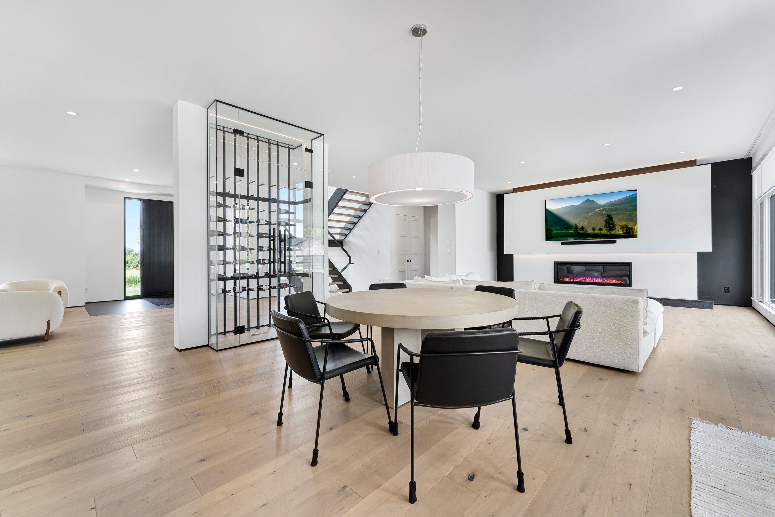 Modern open-plan living room with a round dining table, black chairs, glass wine rack, staircase, white sectional sofa, electric fireplace, and wall-mounted TV. Floor-to-ceiling windows provide natural light.