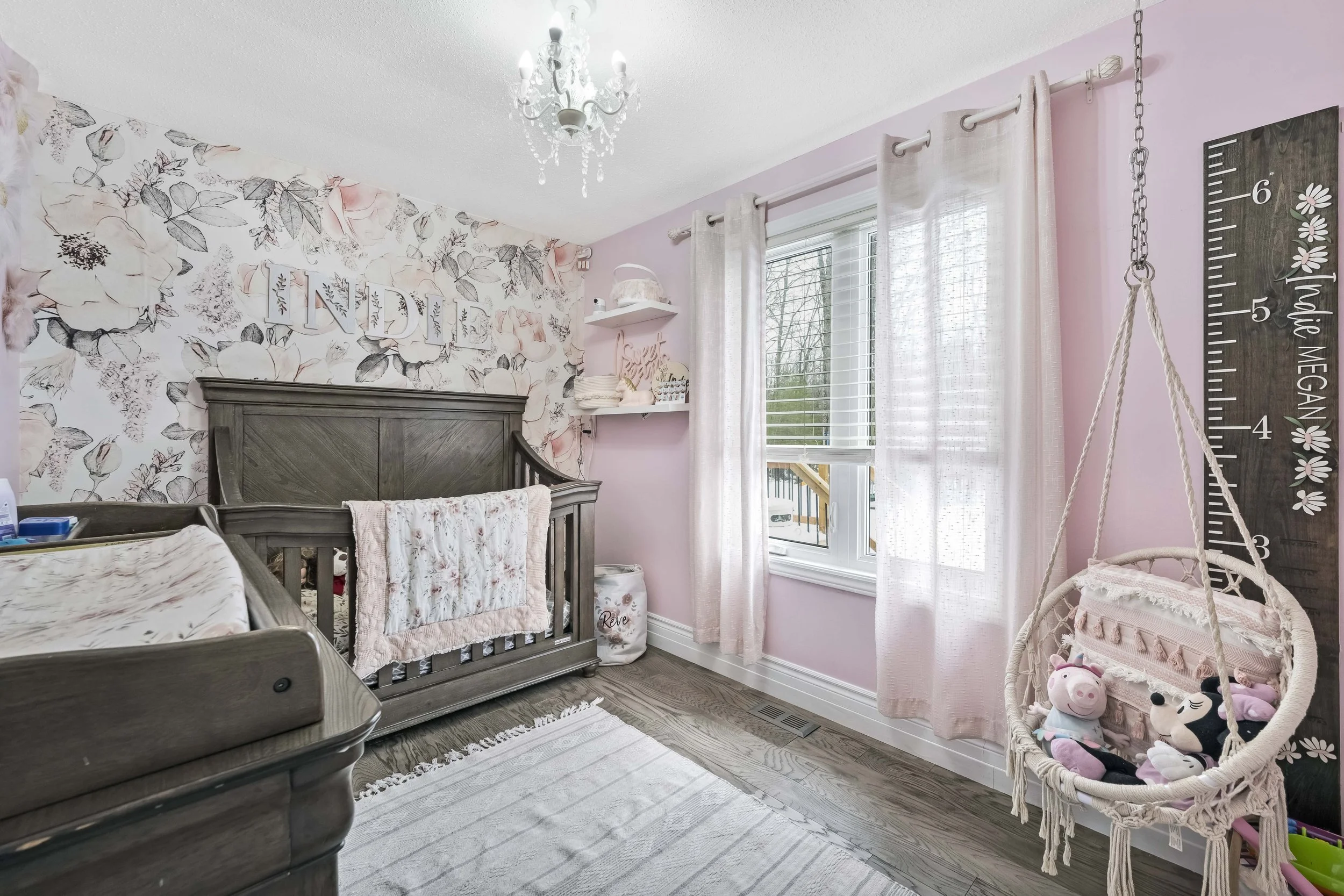 A baby nursery decorated in pink and gray, featuring a gray wooden crib with a pink and white quilt, floral wallpaper with the word 'LINDSEY' above it, shelves with decor items, white curtains, a height measurement wall hanging with plush toys, and a