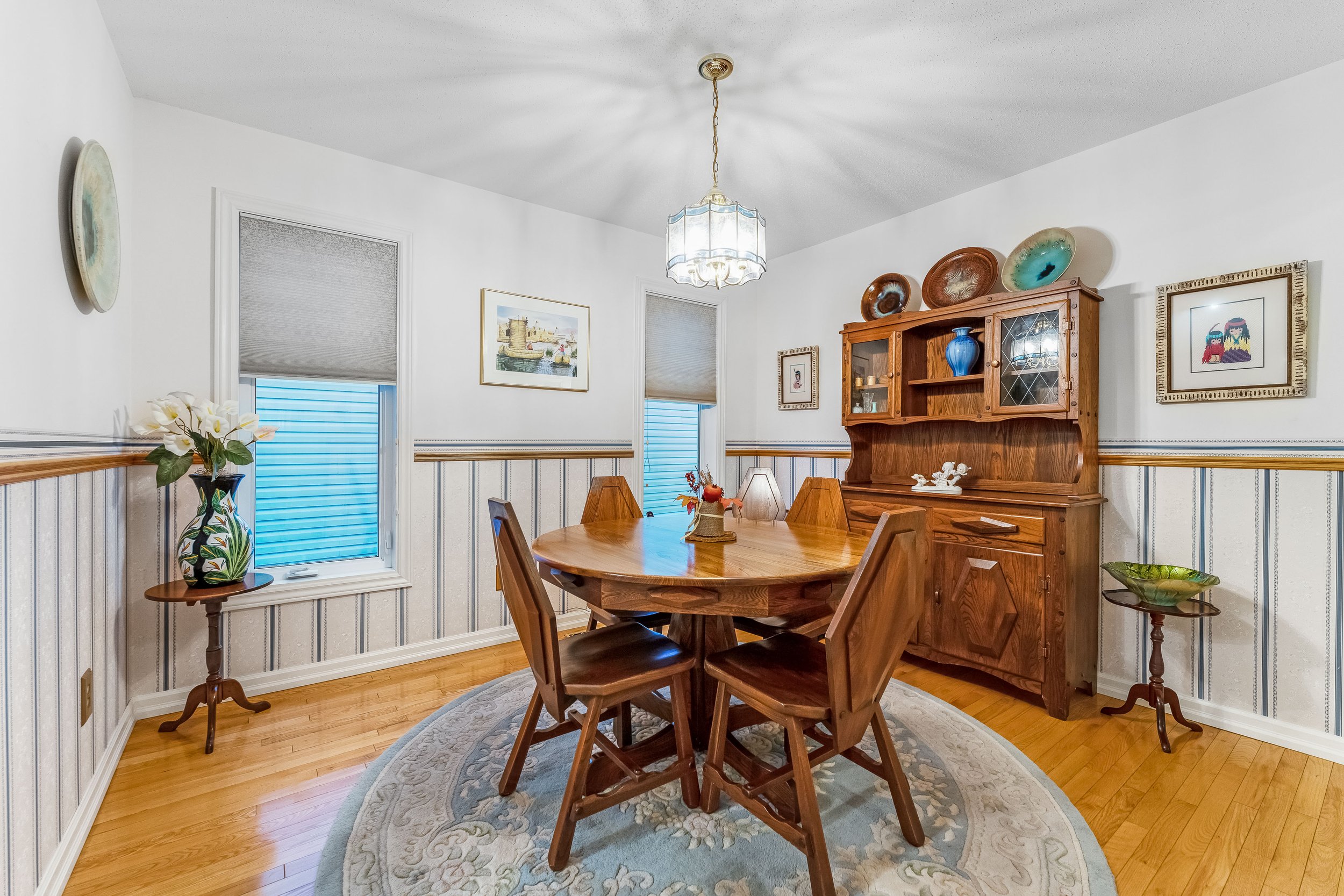 A dining room with a round wooden table and four matching chairs, a wooden hutch with decorative plates and vases, framed artwork on white walls, two windows with gray blinds, a floral rug, and indoor plants.