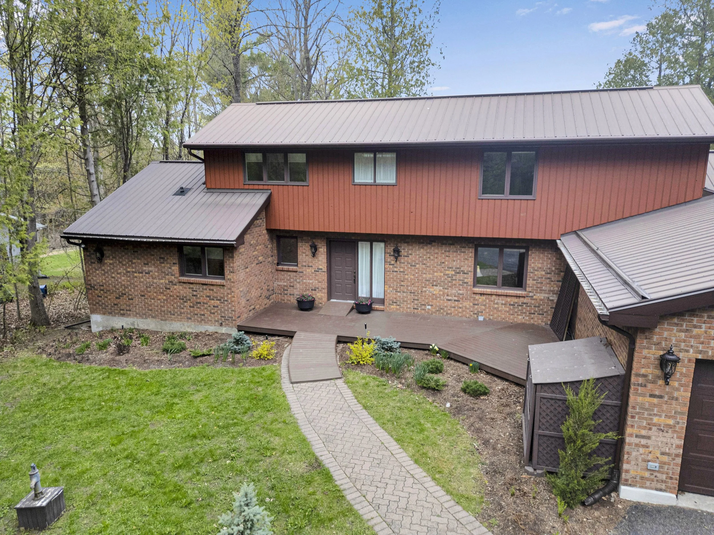 Ottawa Real Estate Media Services  A brick and wood house with a sloped metal roof, surrounded by green grass, plants, and trees with a blue sky overhead.