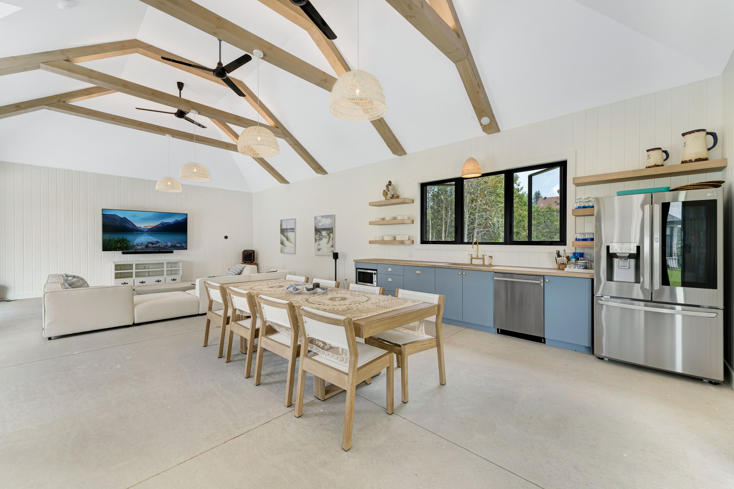 Spacious modern living room with vaulted ceiling and exposed wooden beams. Features a dining area with wooden chairs, a large flat-screen TV, cream-colored sofas, and a kitchen section with blue cabinets and stainless steel appliances.