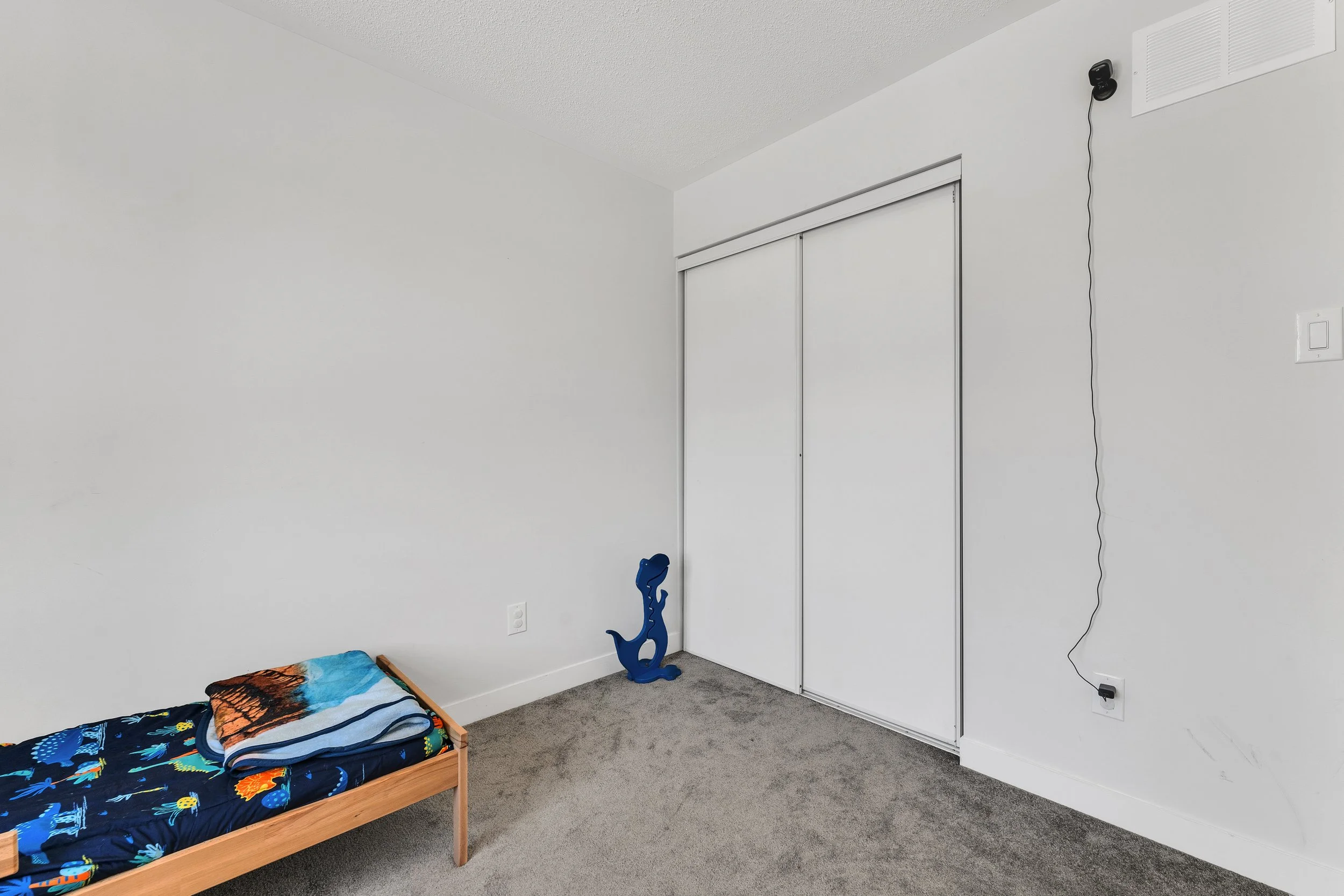 NV3D Real Estate Photography Ottawa  Empty bedroom with a small bed with colorful bedding, a blue toy dinosaur, and a white closet with sliding doors.