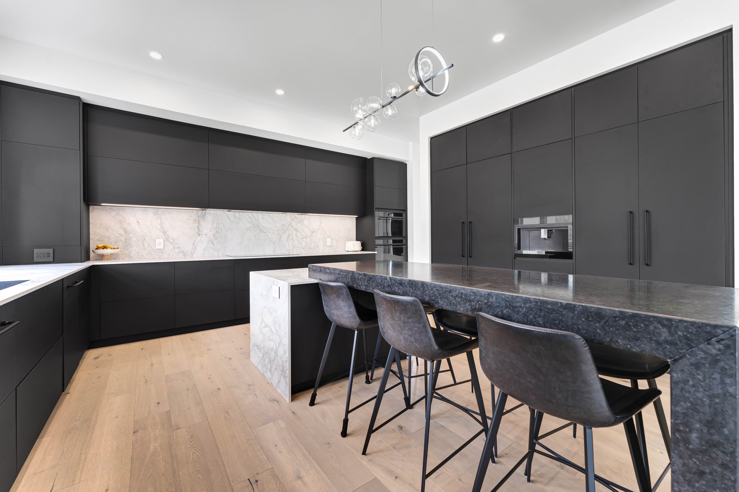 Modern kitchen with black cabinets, marble countertops, kitchen island with bar stools, and minimalist lighting fixture.