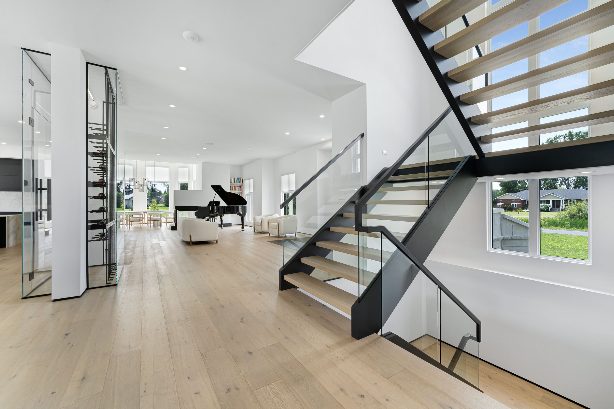 Modern interior with open space design, wooden floors, glass staircase, a grand piano, wine rack, and large windows providing natural light.