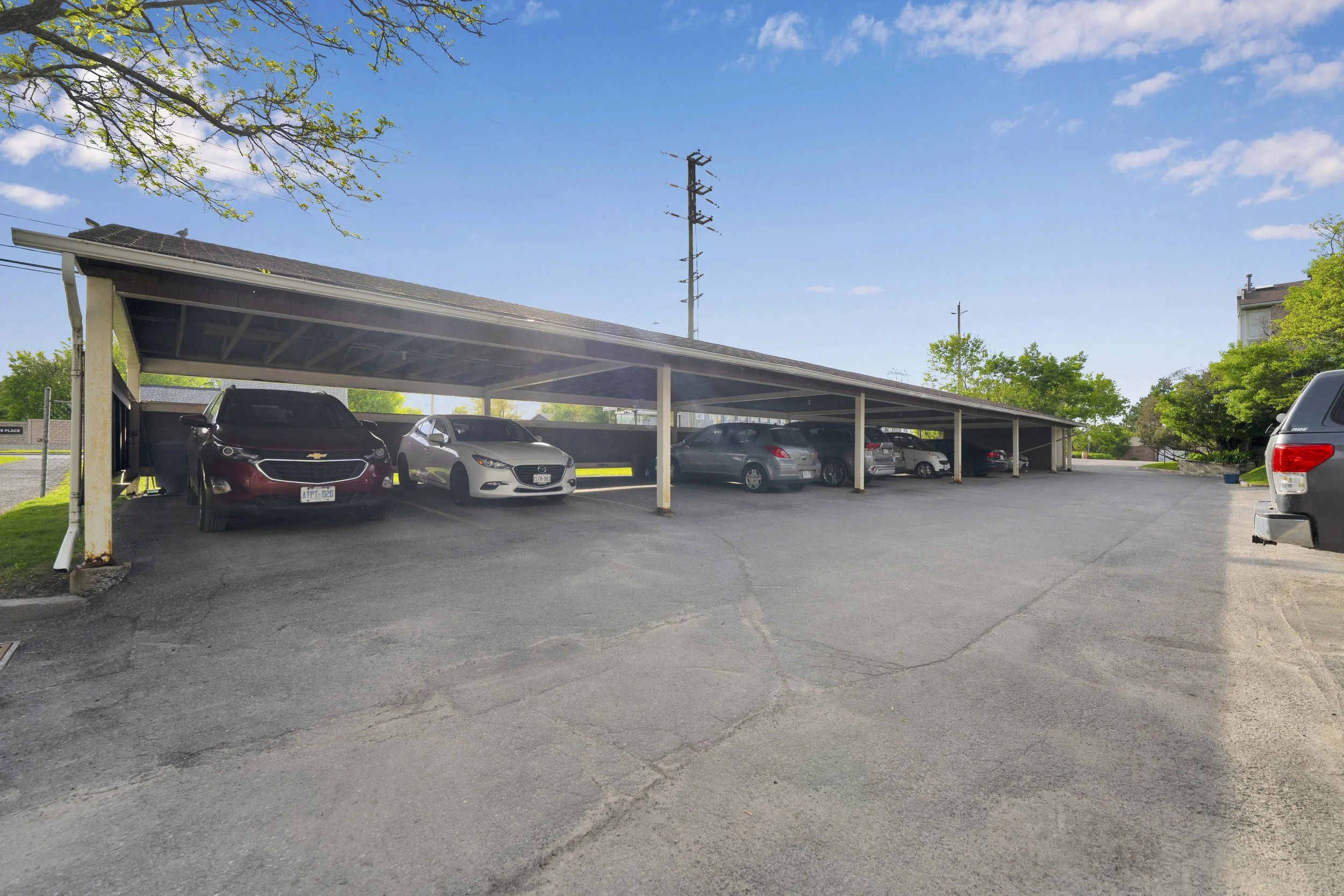 NVISION3D Real Estate Photography Ottawa  A parking lot with covered parking spaces, multiple cars parked, and trees with green leaves under a blue sky with some clouds.