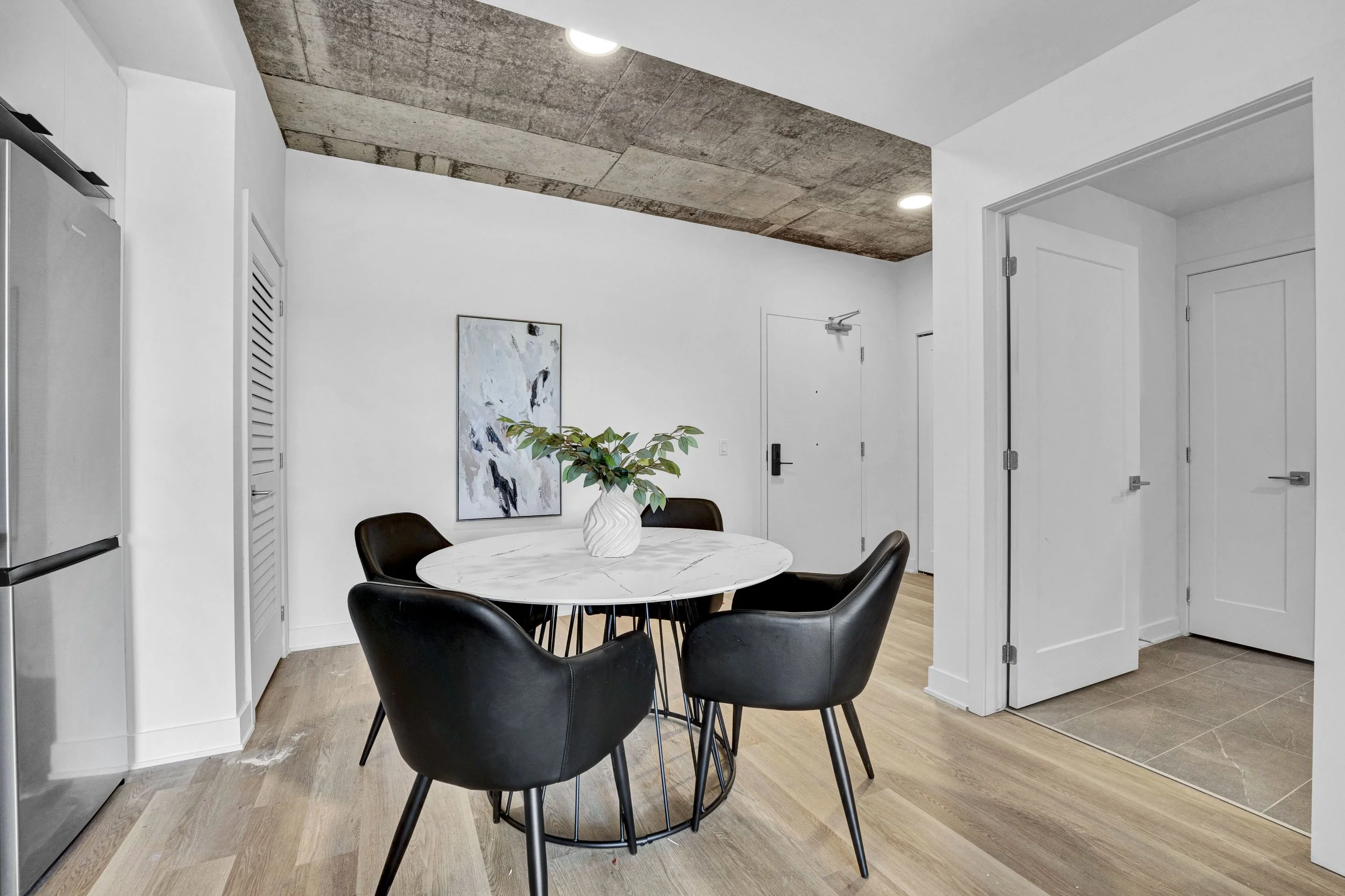 Modern dining room with a round white marble table, black chairs, a white vase with green foliage, white walls, artwork, and wood flooring under a concrete ceiling.