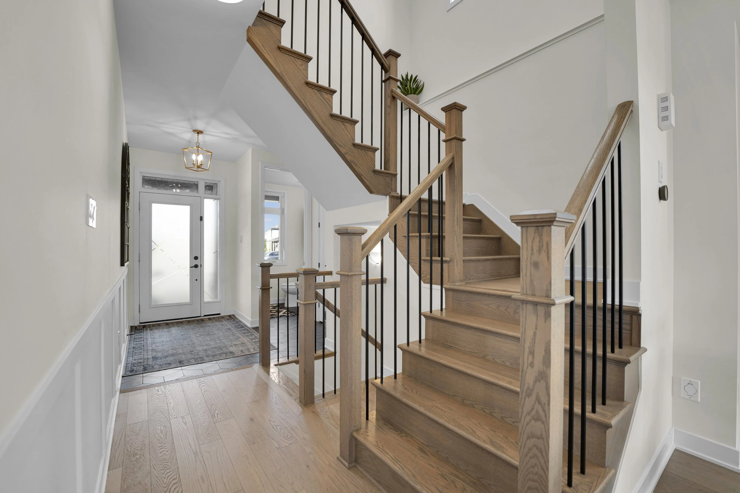 NVISION3D Real Estate Photography Ottawa  Interior view of a modern home entryway featuring a wooden staircase with black metal balusters and a light wood floor, with a white front door and a window.