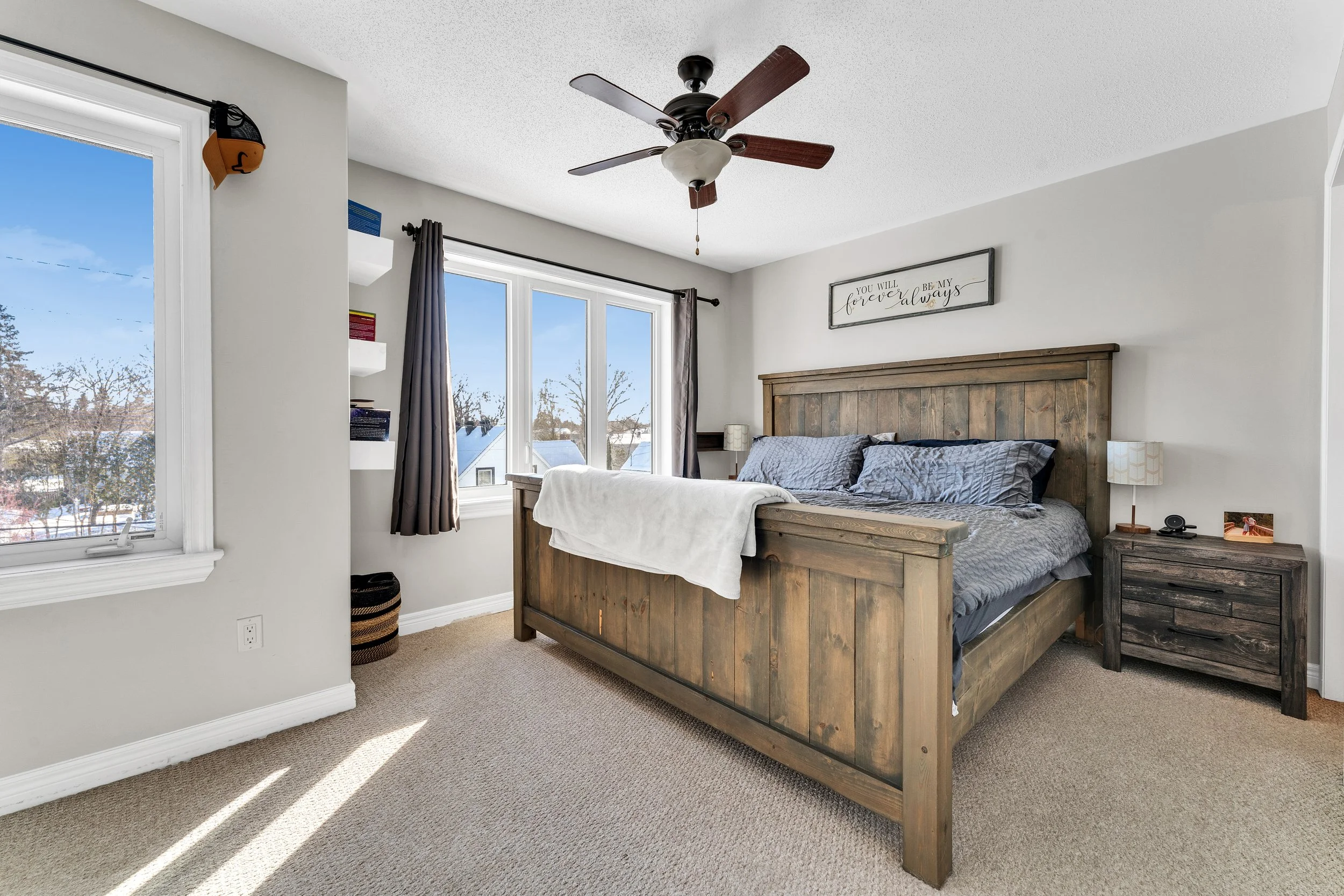 Ottawa Real Estate Photography  A bedroom with a wooden bed, nightstand, ceiling fan, and large windows showing a snowy outdoor scene.