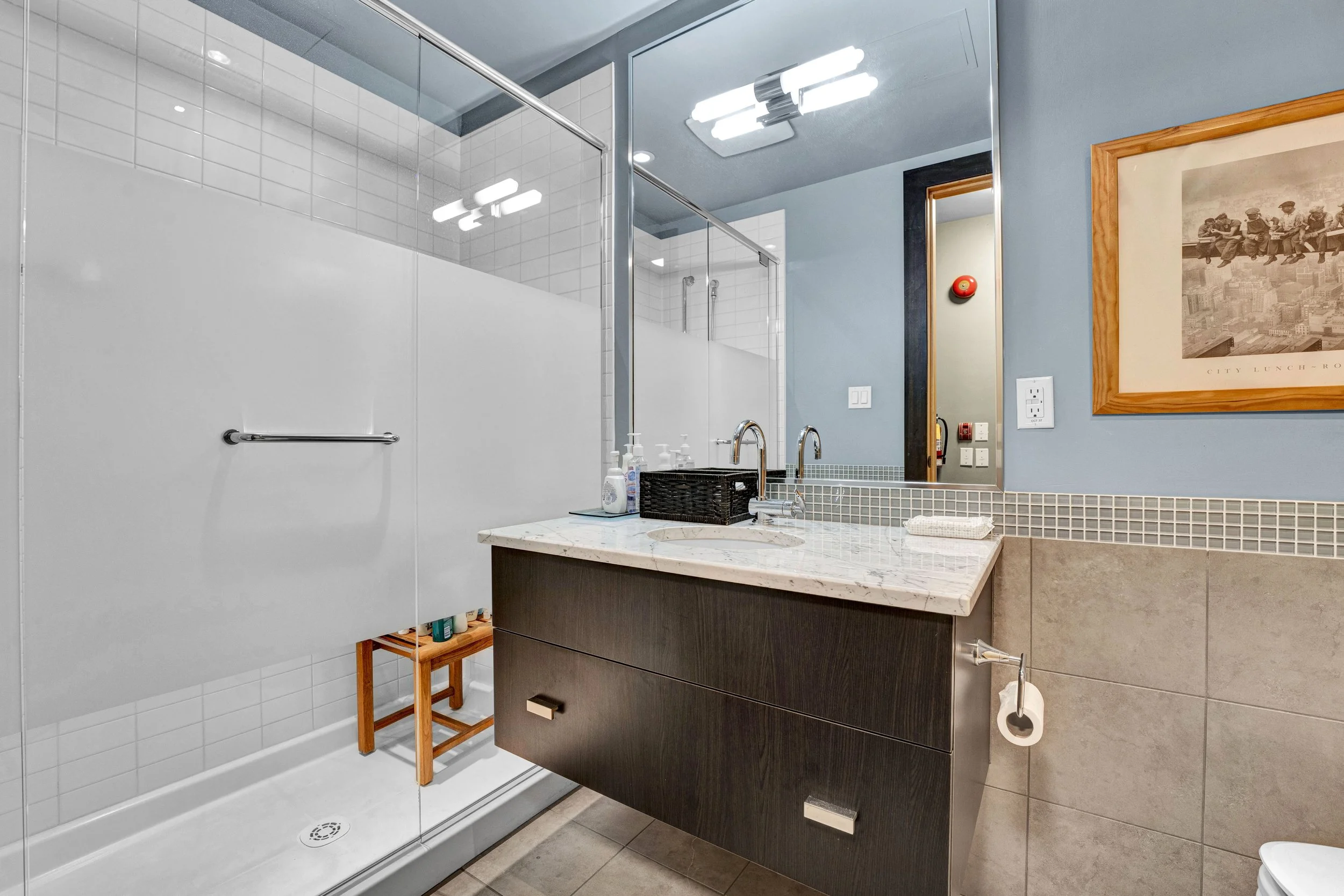 Ottawa Real Estate Photographer NVISION3D.ca  Modern bathroom with a glass enclosed shower, a dark wood vanity with a white marble countertop, a large mirror, and a framed black and white cityscape photograph on the blue wall.