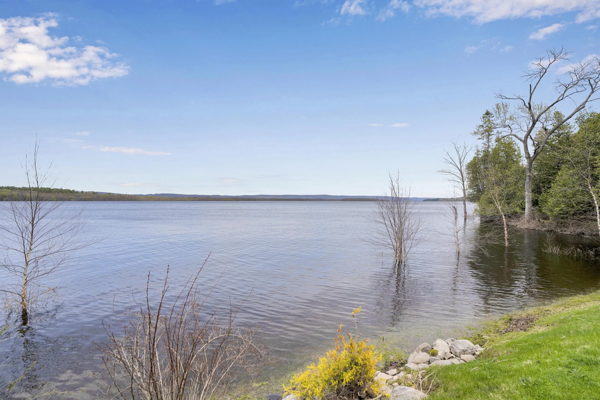 NVISION3D.ca Real Estate Media Ottawa  A landscape of a large body of water, possibly a lake or river, with a tree-covered shoreline on the right and a few leafless trees in the foreground. The sky is mostly clear with some scattered clouds.
