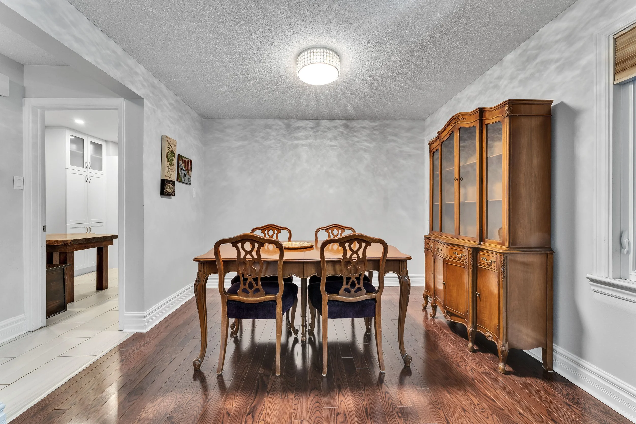 Ottawa Real Estate Photography NVISION 3D Dining room with a wooden table, four chairs, and a wooden china cabinet against the wall. There are two pieces of wall art on the left wall, and a doorway leading to a kitchen area.