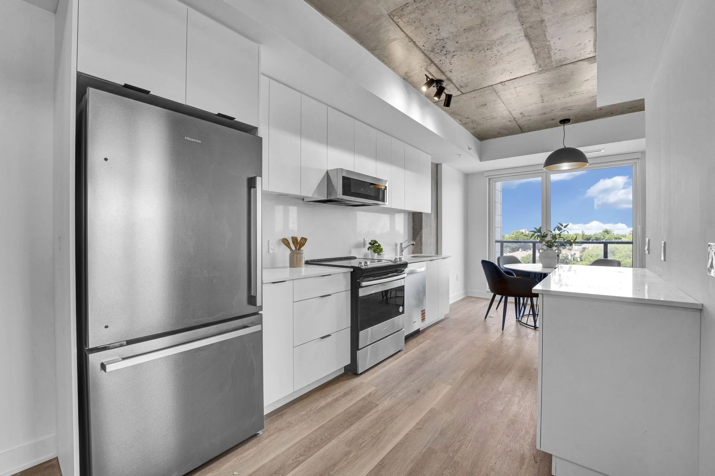 Modern kitchen with white cabinets, stainless steel refrigerator, microwave above stove, and large window with a city view.