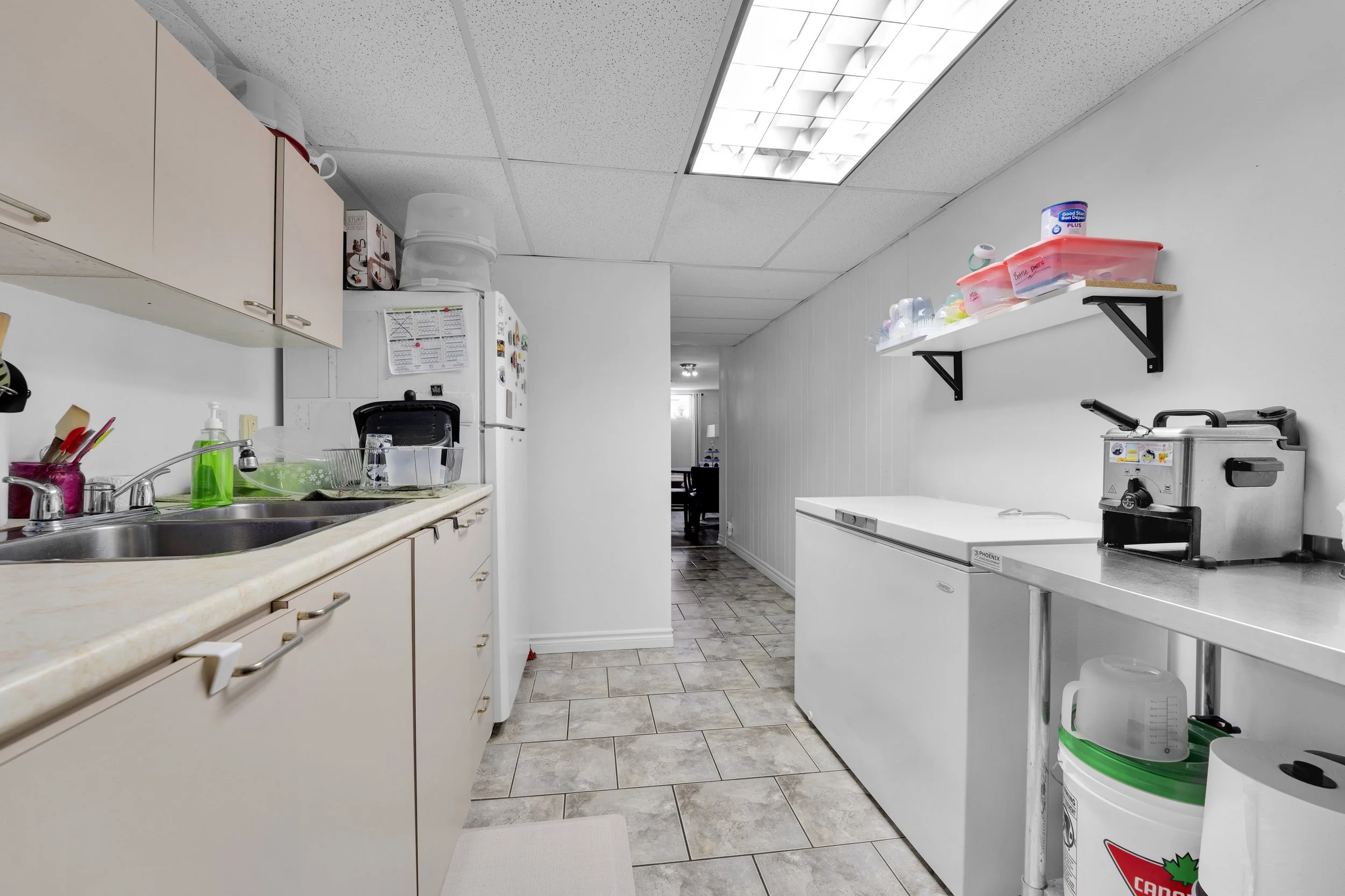 NVISION3D.ca Real Estate A kitchenette with countertops, cabinets, a sink with soap, a dish rack, a refrigerator, a microwave, shelves with containers, a sterilizer, and a trash can in a room with tiled flooring and a ceiling with fluorescent lights.