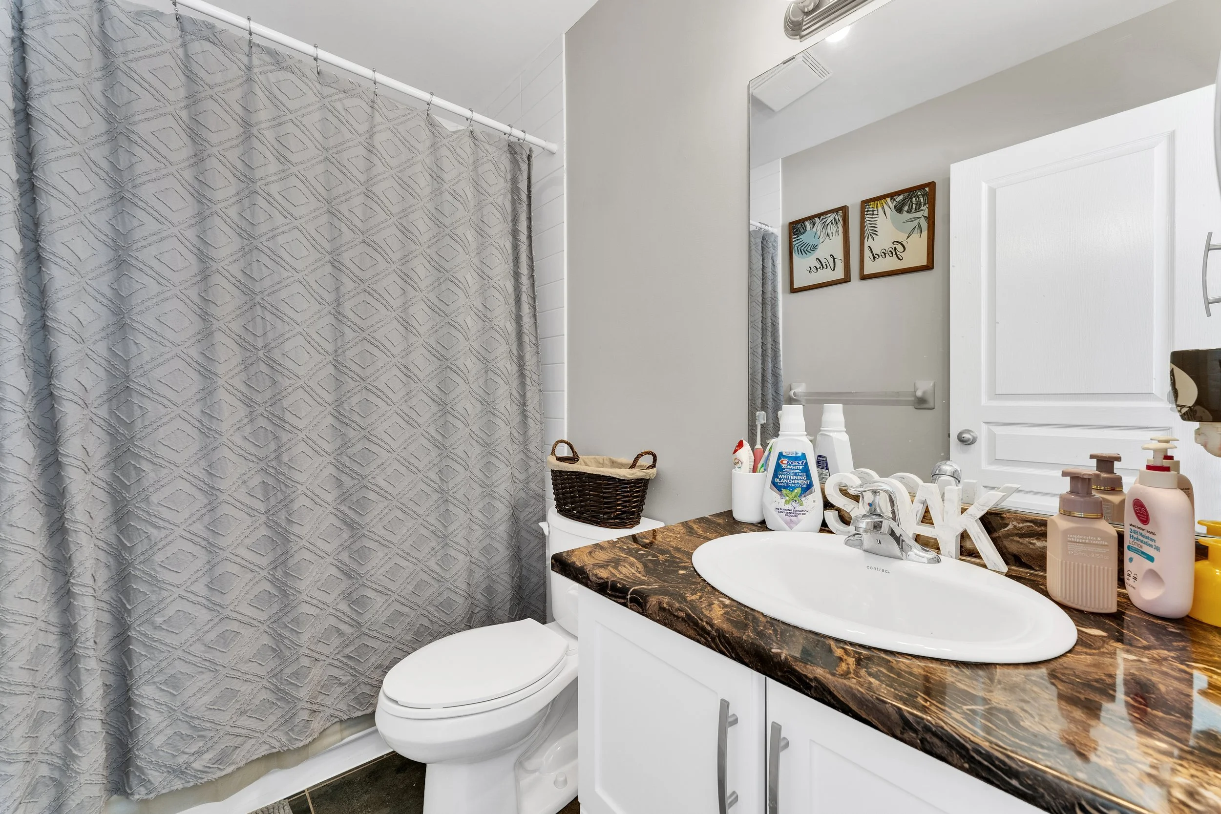Bathroom with white vanity, marble countertop, oval sink, large mirror, framed wall art, shower with textured curtain, and various toiletries.