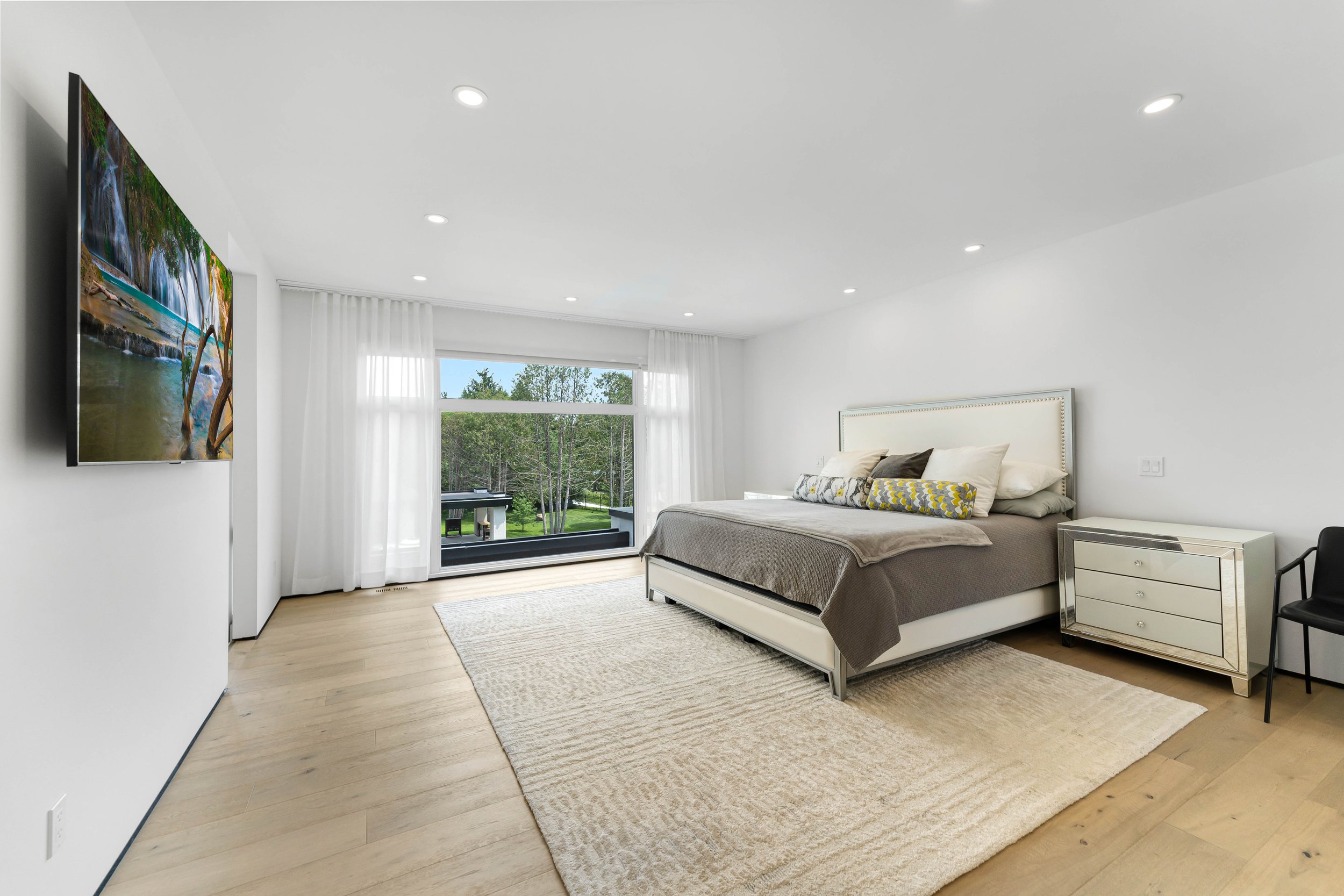 Modern bedroom with a large bed, white and gray bedding, a mirrored nightstand, and a wall-mounted flat screen TV. A large window with sheer drapes offers a view of trees outside. The room features recessed lighting and light wood flooring with a bei