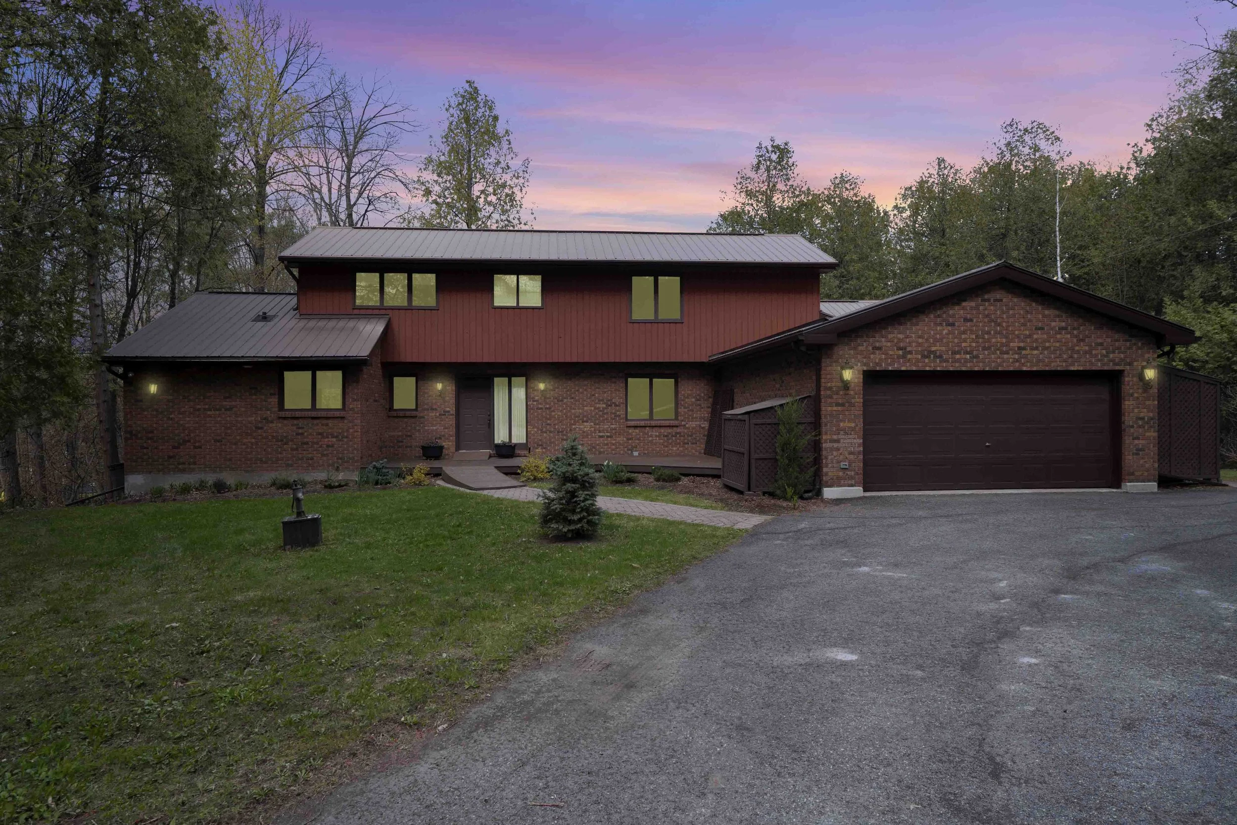 Ottawa Real Estate Media Services  A two-story brick house with a metal roof, situated on a grassy lawn with young trees and shrubs, at dusk with a colorful pink and purple sky.