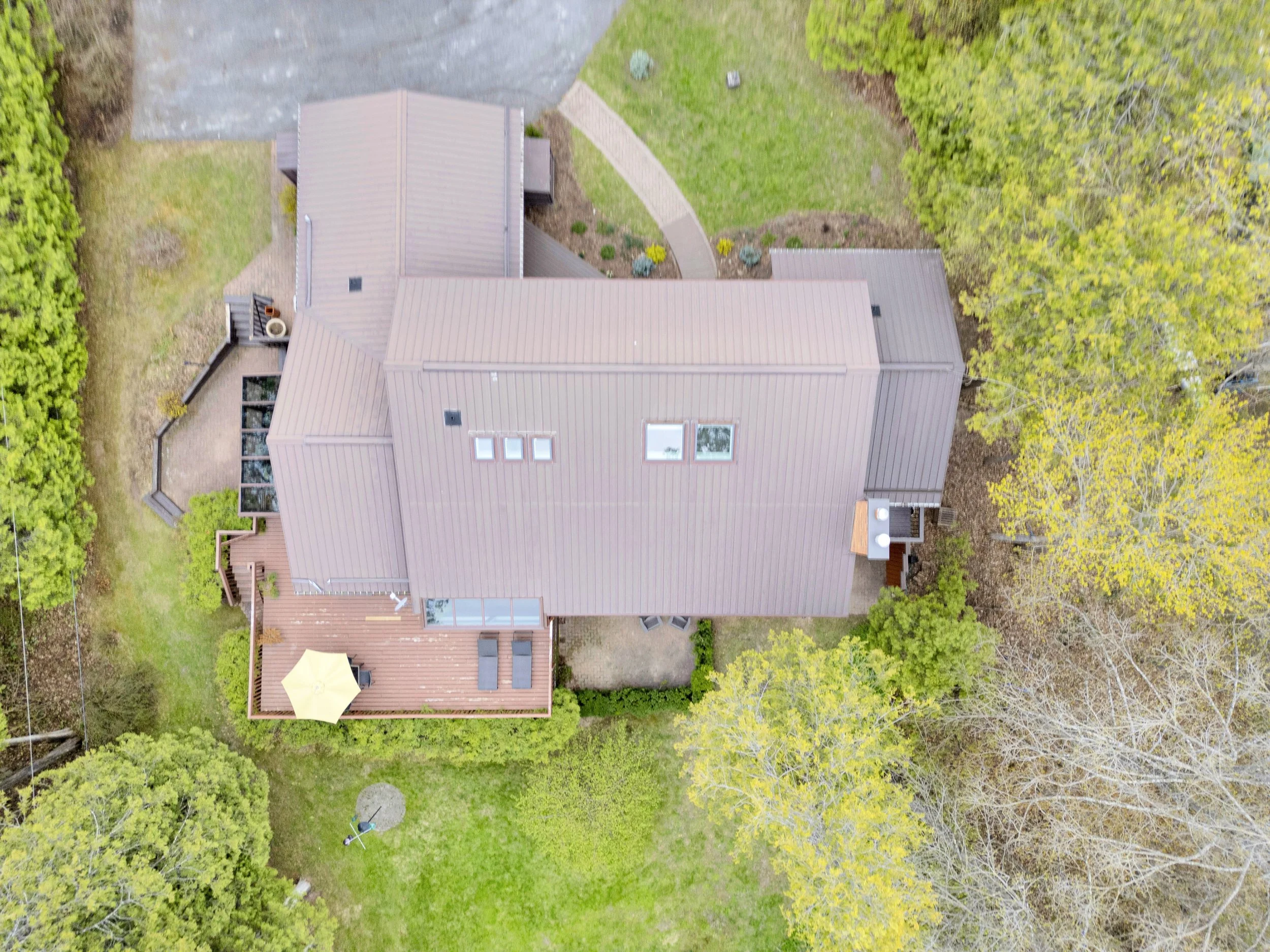 Ottawa Real Estate Media Services  An aerial view of a backyard showing a house with a brown roof, a patio with a yellow umbrella, a lawn, trees surrounding the yard, and a paved driveway.