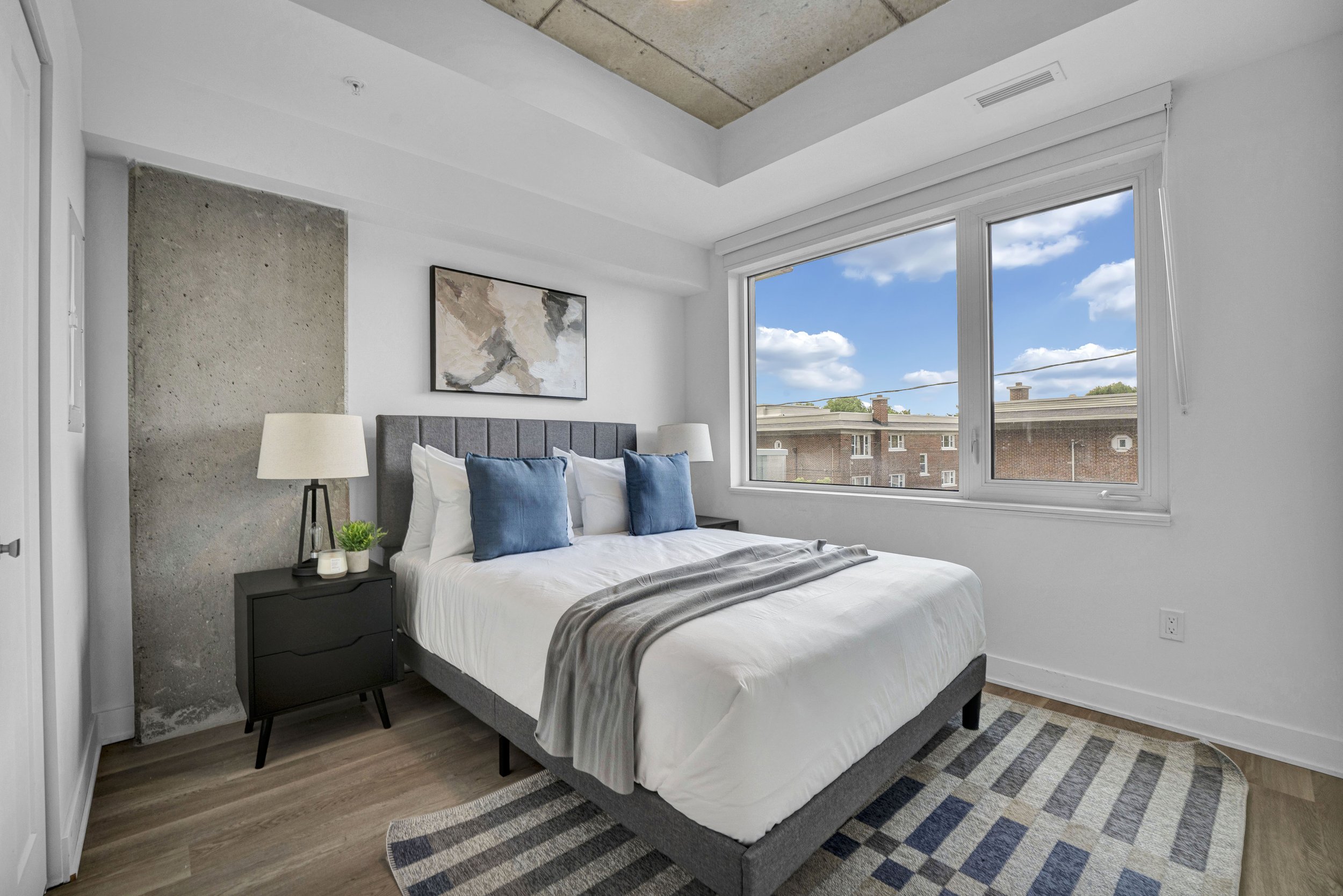 A modern bedroom with a gray upholstered bed, white bedding, two blue pillows, a black nightstand with a lamp, small plants, a framed abstract artwork on the white wall, large window with a view of neighboring buildings and blue sky with clouds, and 
