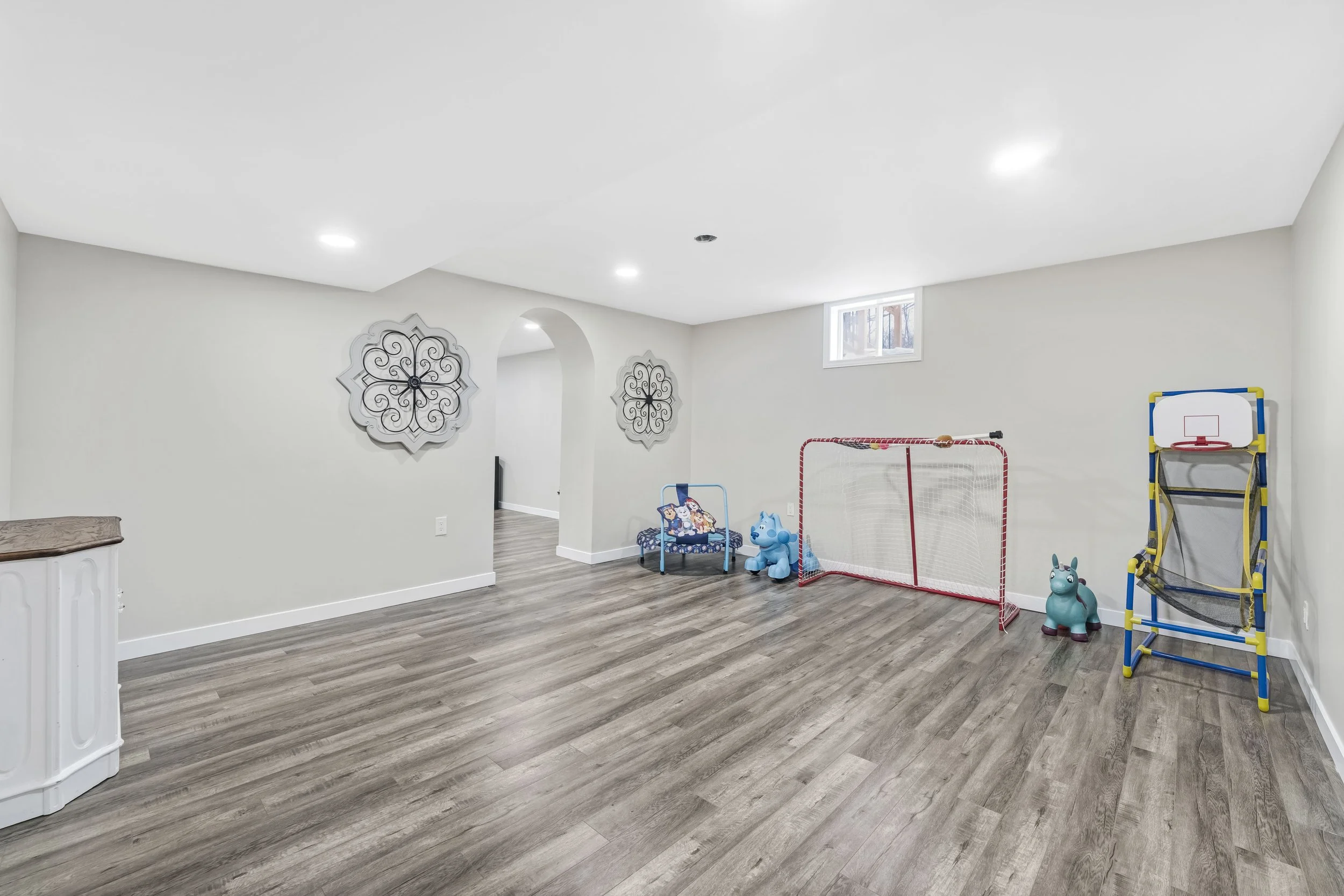 Empty basement room with white walls, wood floor, ceiling lights, and children's toys including a small basketball hoop, a small chair, a blue hippo, and a playpen.