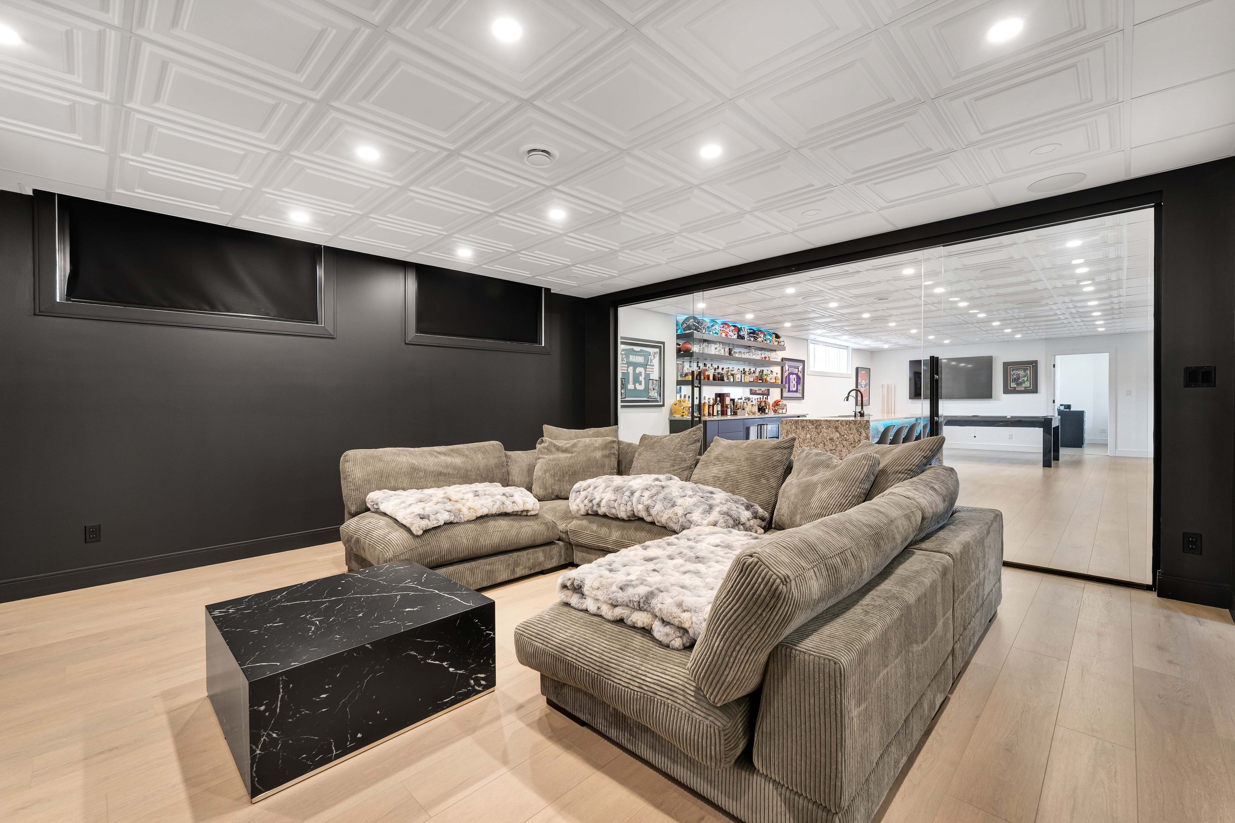 Modern home theater with large sectional sofa, cozy blankets, black marble coffee table, and view of an adjacent game room with sports memorabilia, bar, and pool table.